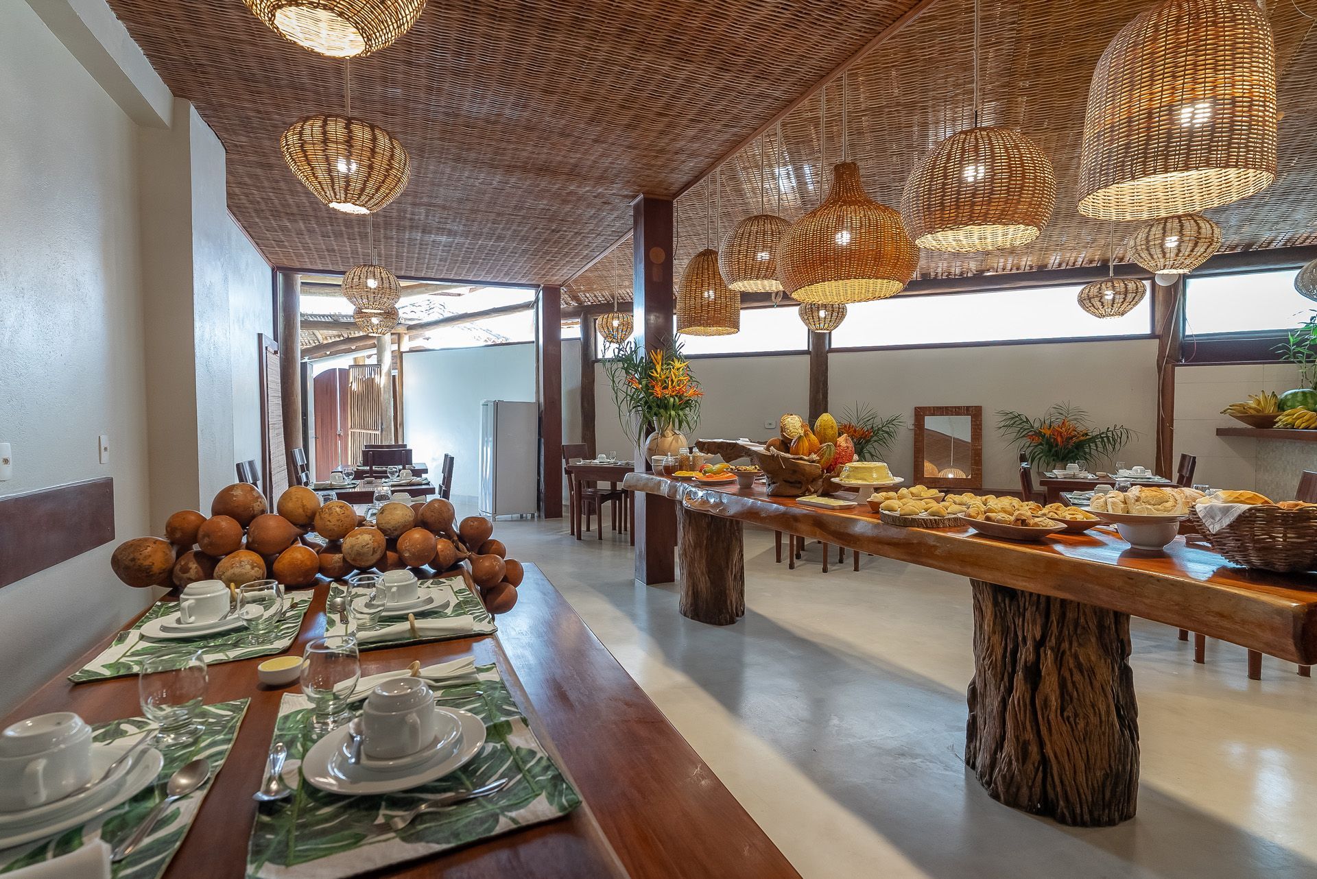 Buffet de café da manhã em uma sala de jantar rústica com mesas de madeira, luminárias de vime e uma variedade de alimentos.
