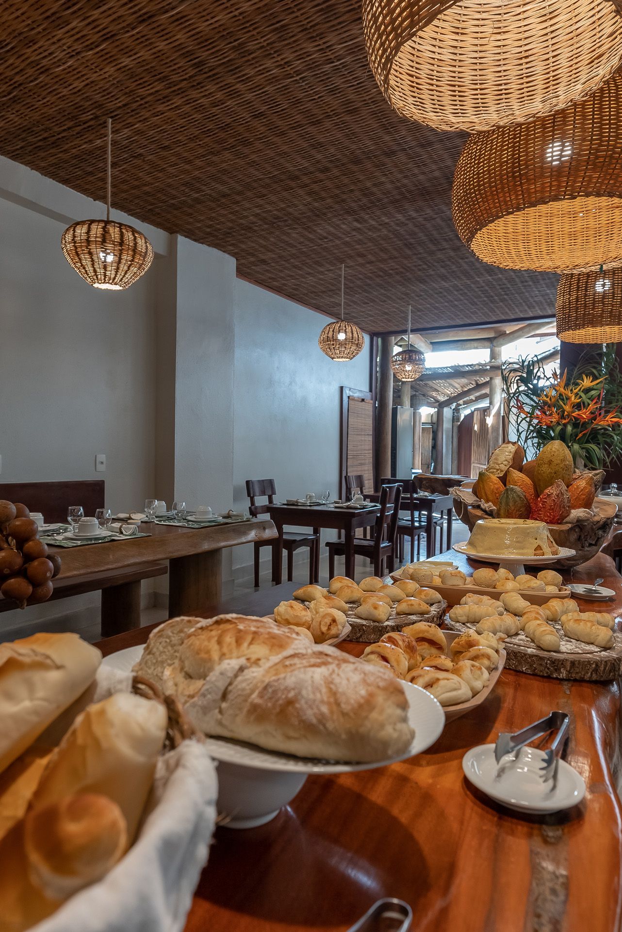Buffet de café da manhã com diversos tipos de pães, doces e uma longa mesa de madeira posta para os comensais, sob luminárias de vime.