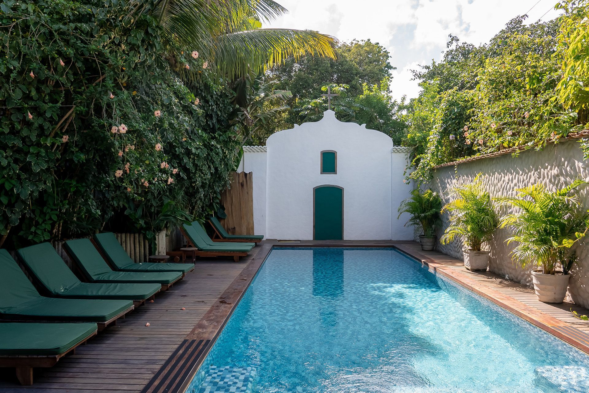 Piscina rodeada de vegetação, com espreguiçadeiras e estrutura branca com porta verde.