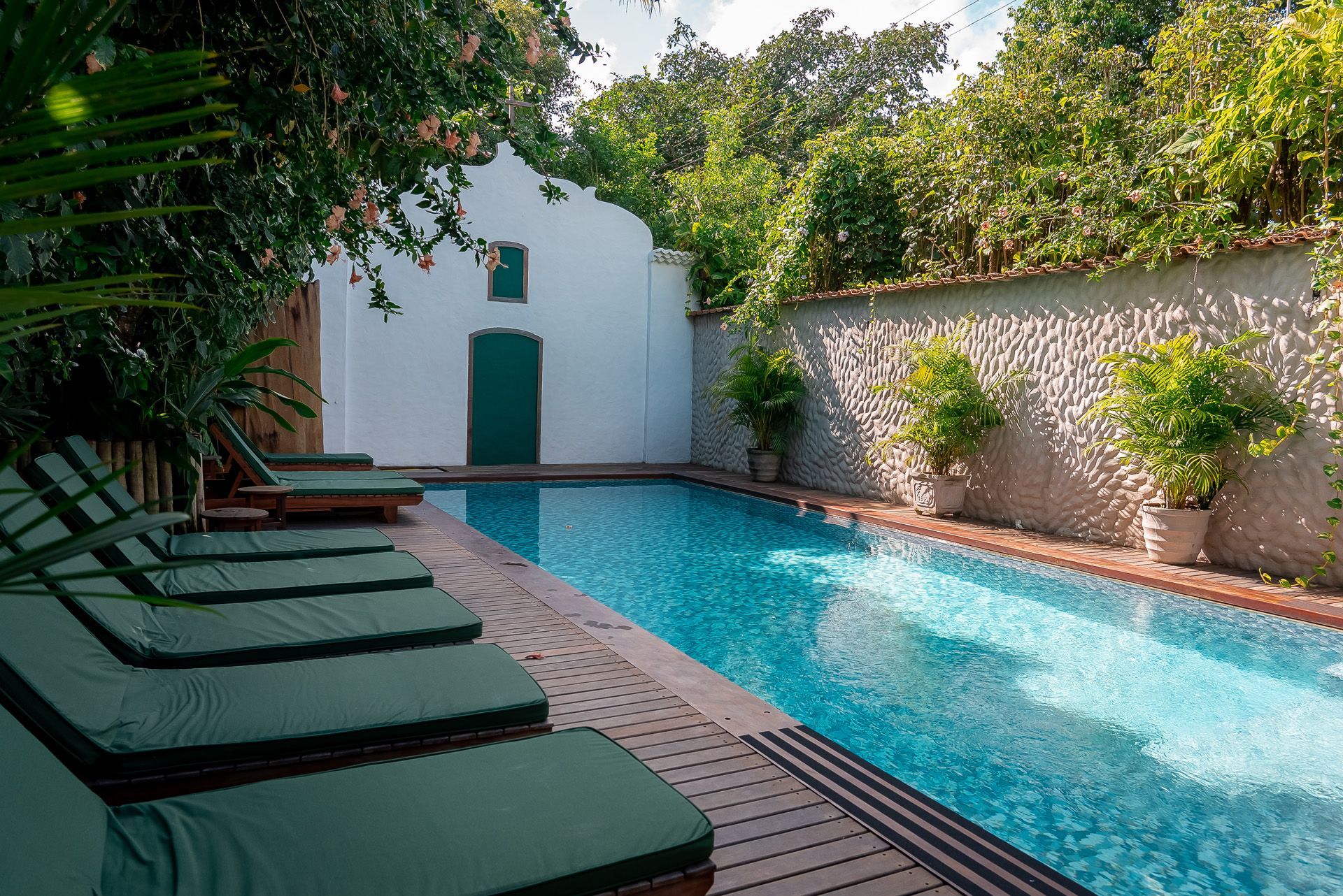 Piscina com espreguiçadeiras ao lado de um prédio branco com uma porta verde; vegetação exuberante ao redor.