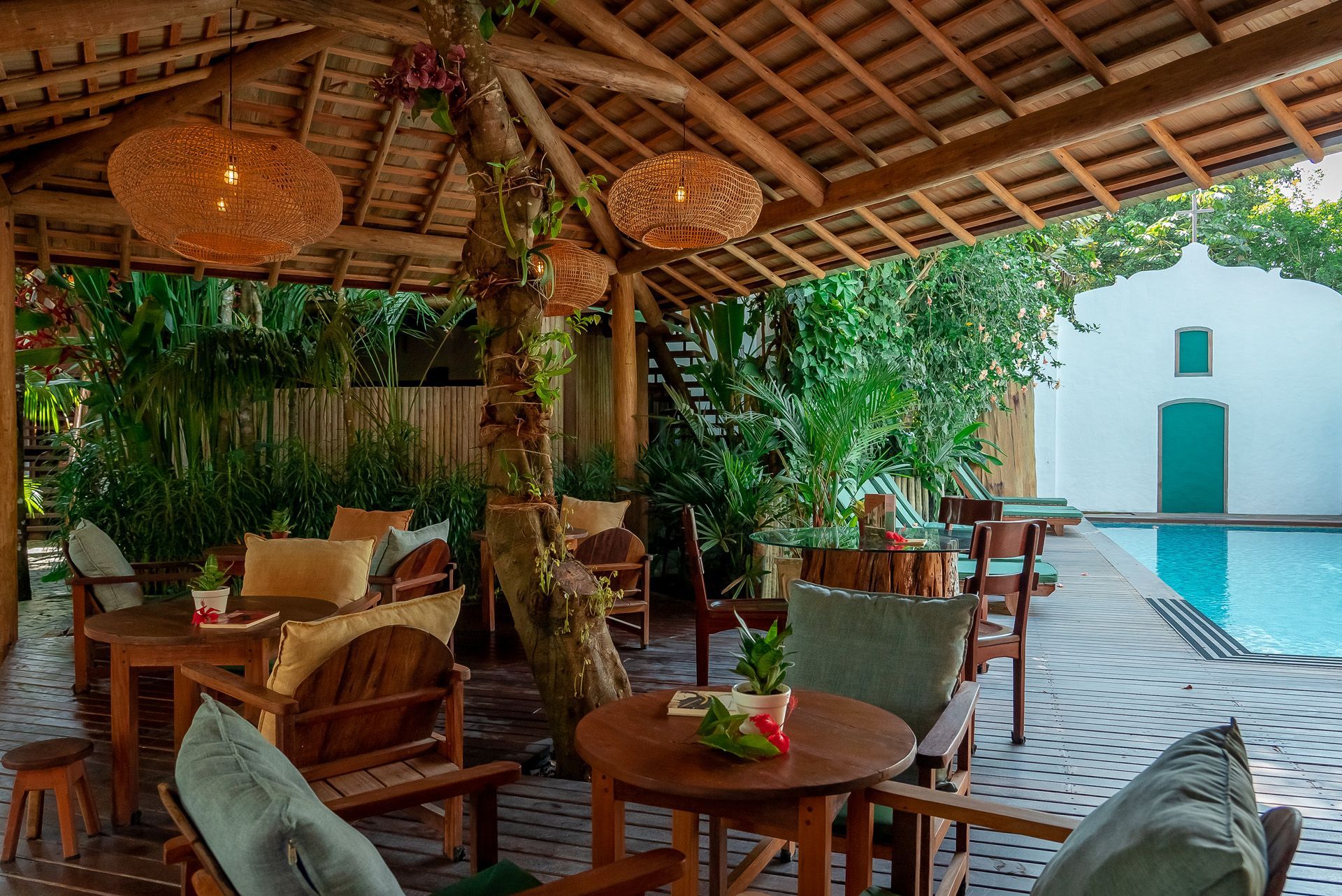 Área de estar com móveis de madeira, luminárias de vime e piscina. Vegetação exuberante e um prédio branco ao fundo.