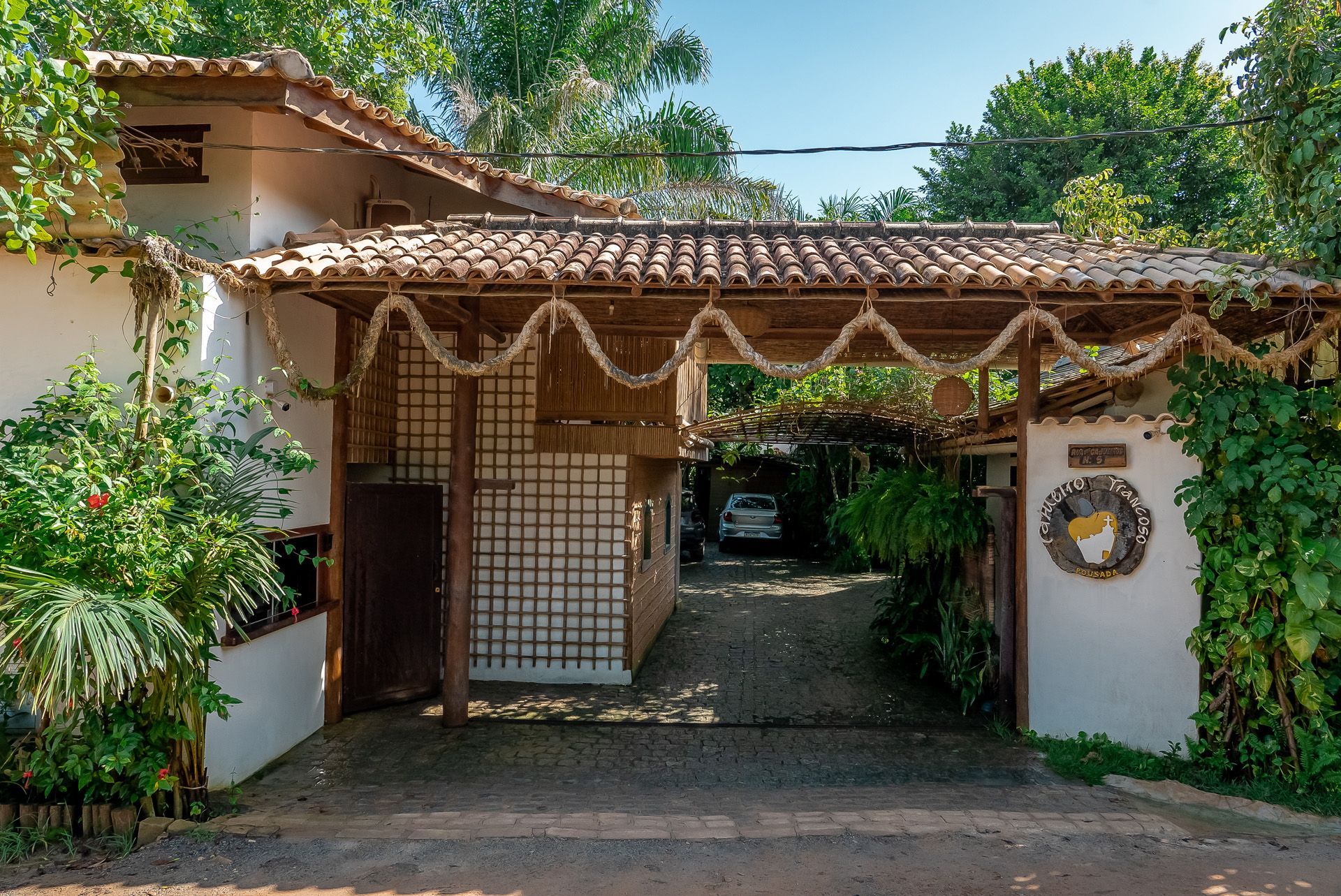Entrada de um edifício com telhado de telhas, aberta para uma garagem com um carro estacionado. Vegetação exuberante circunda a estrutura.