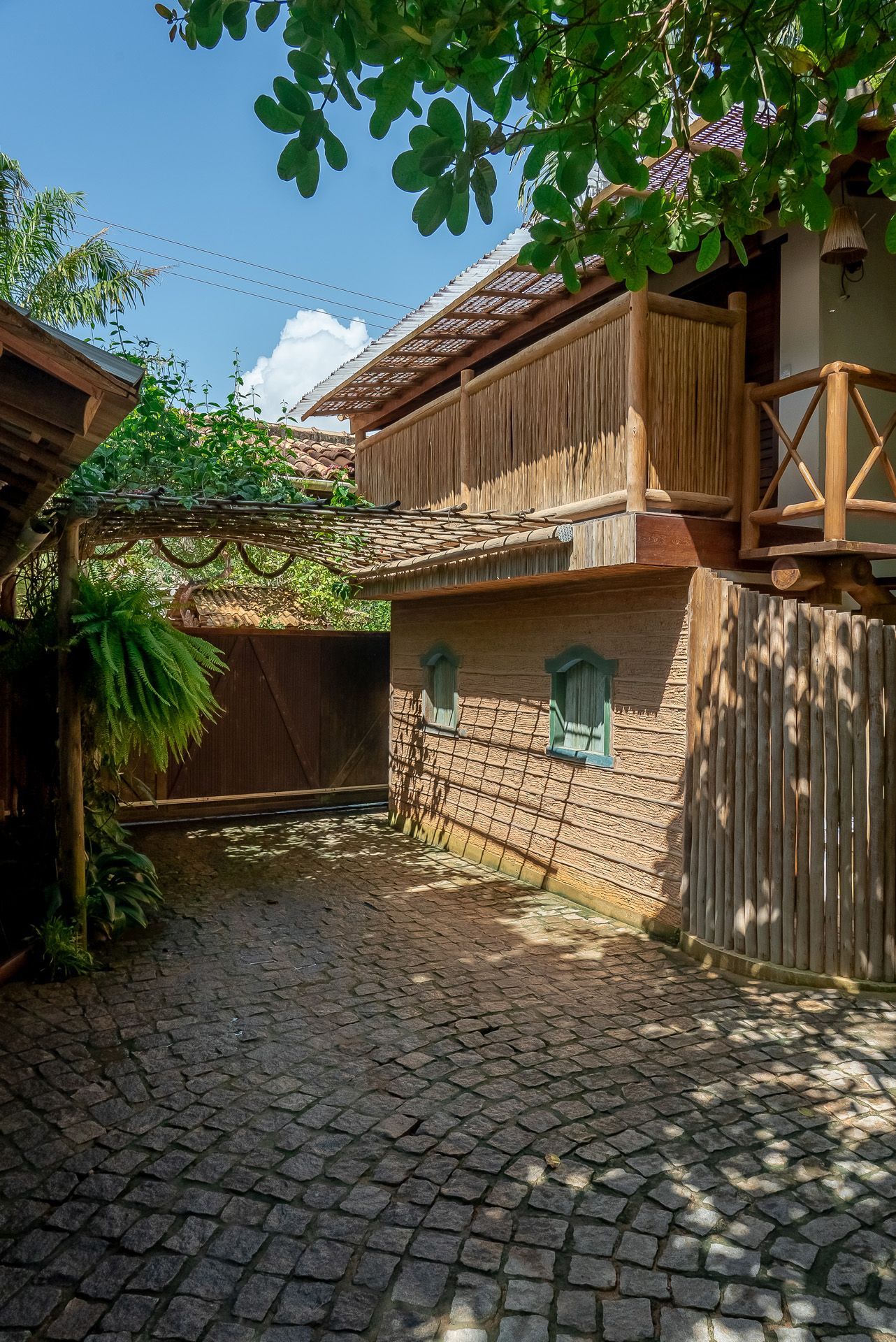 Um caminho de pedra conduz a uma construção rústica com varanda e portão de madeira, sob uma folhagem exuberante.