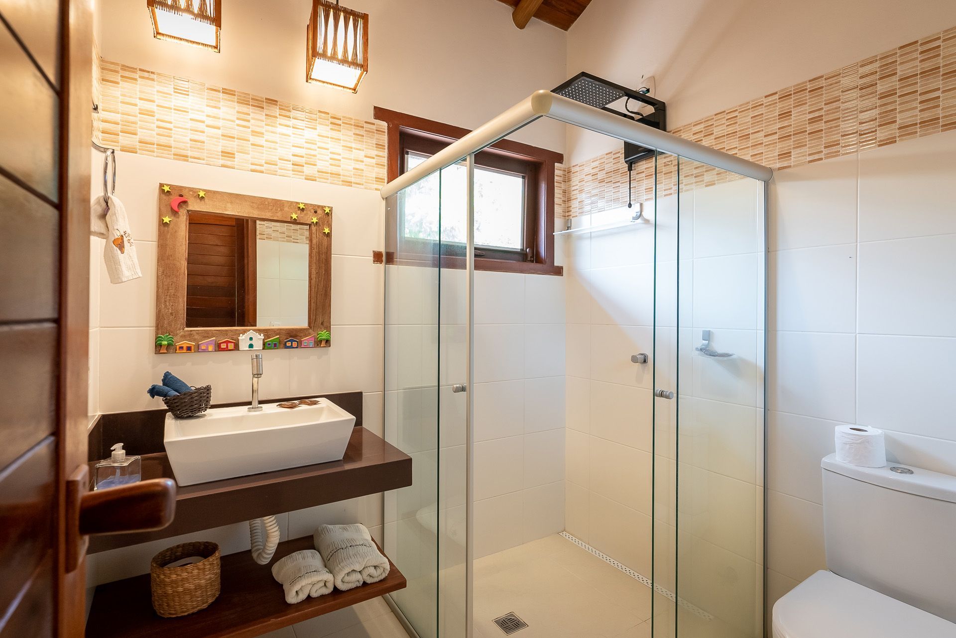 Banheiro com box de vidro, pia e vaso sanitário. Azulejos em tons terrosos e detalhes em madeira.