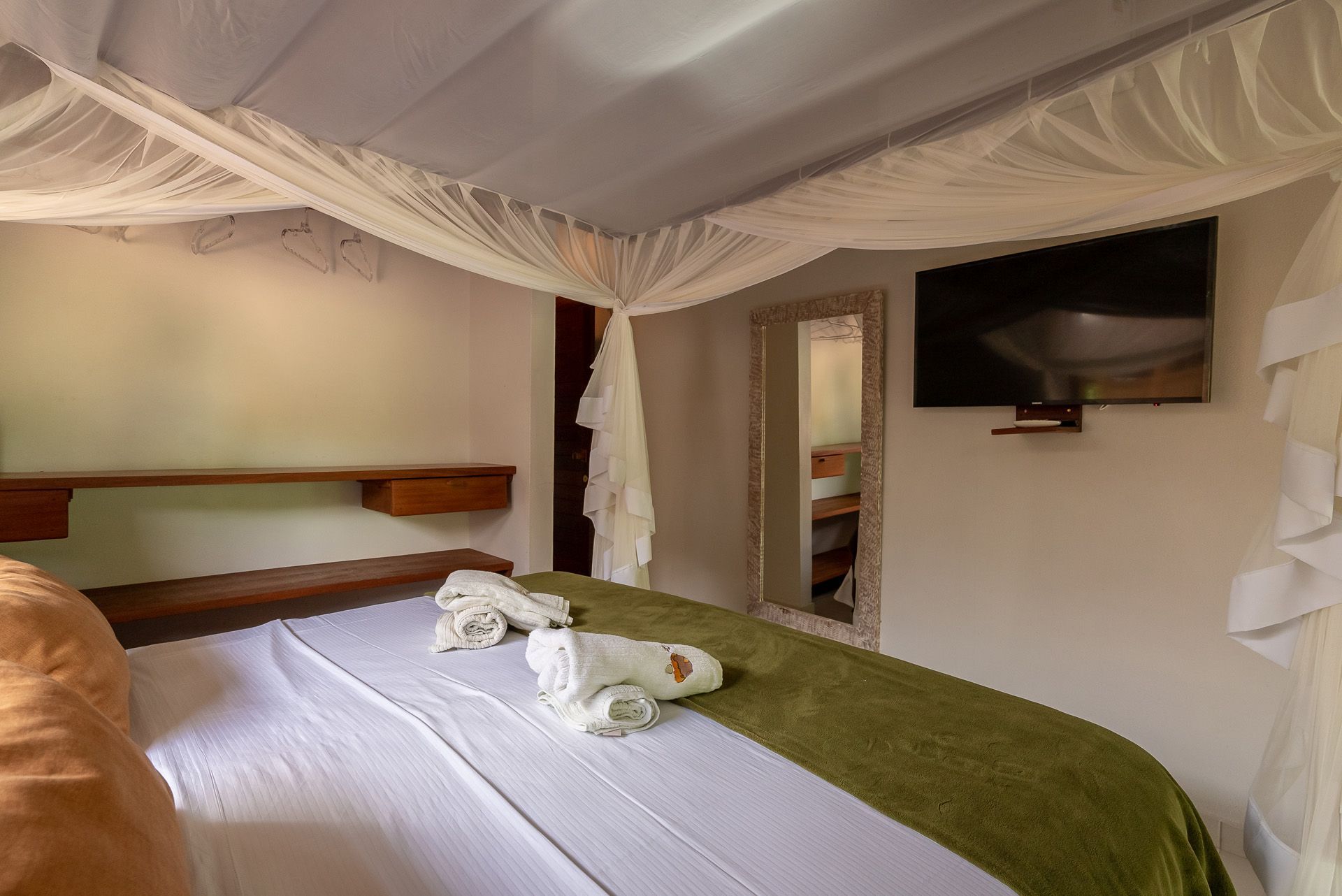 Quarto com cama branca, tapete verde, mosquiteiro, espelho e TV fixada na parede.