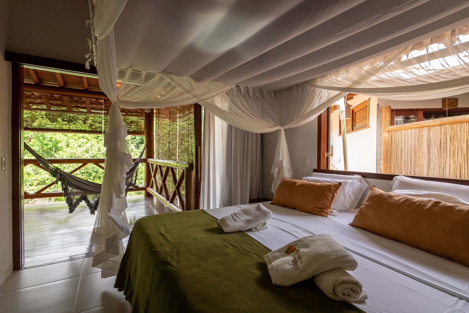 Quarto com cama de dossel, varanda com vista para vegetação exuberante, rede e luz natural.