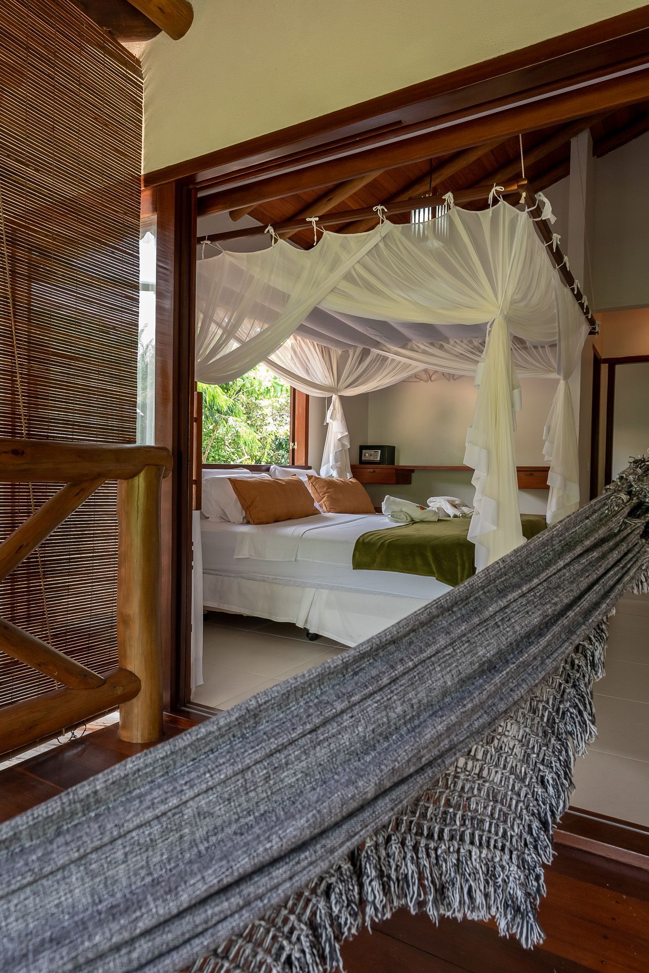 Quarto com rede na varanda, cama com mosquiteiro, acabamento em madeira natural e vegetação visível pelas janelas.