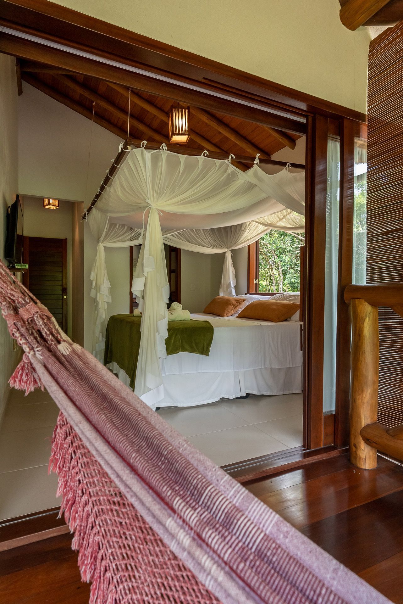 Quarto com porta aberta: cama com dossel, rede, vigas de madeira, luz natural.