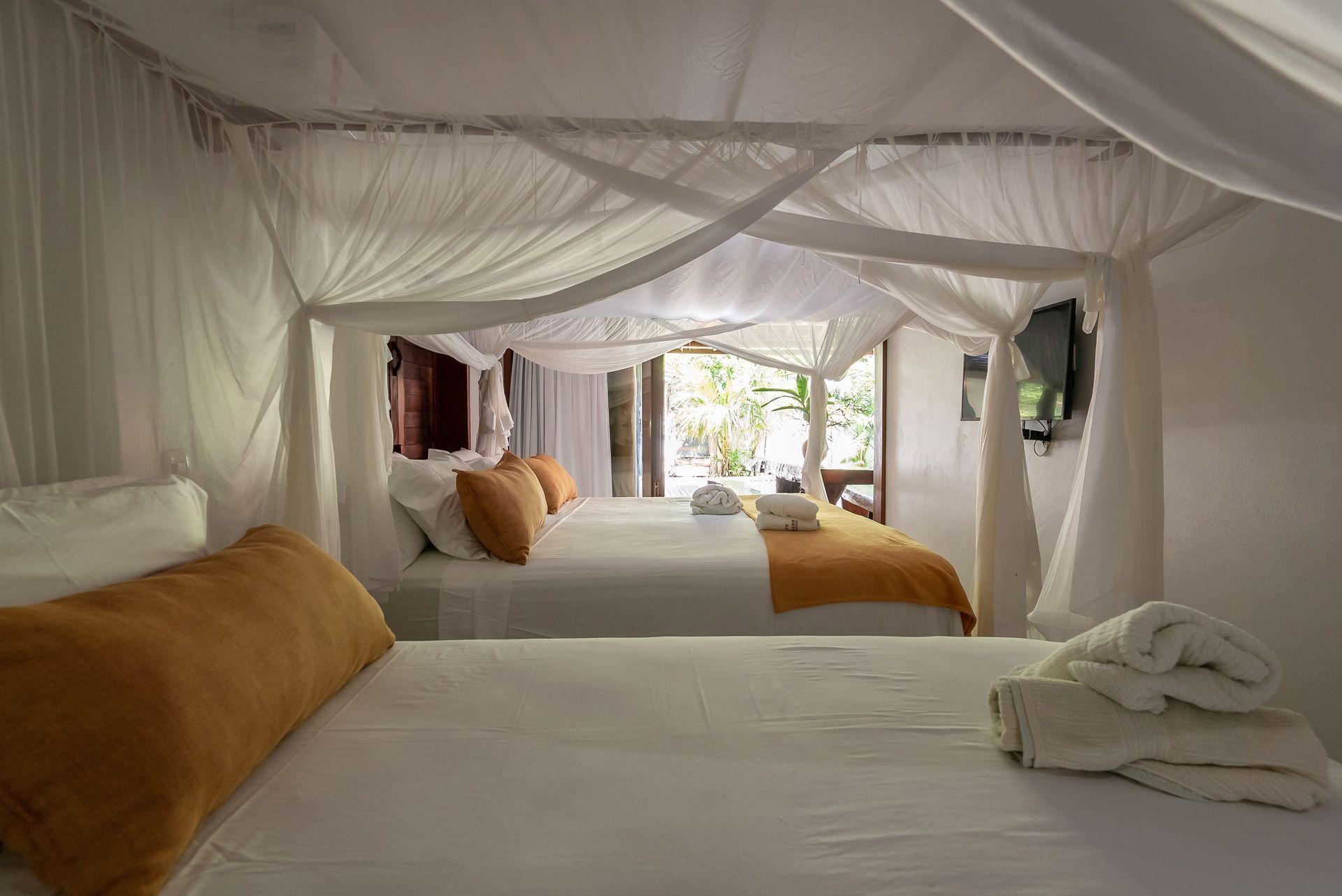 Quarto com cama de dossel branca, duas camas, travesseiros laranja e TV. Quarto amplo e iluminado com porta aberta para um pátio.