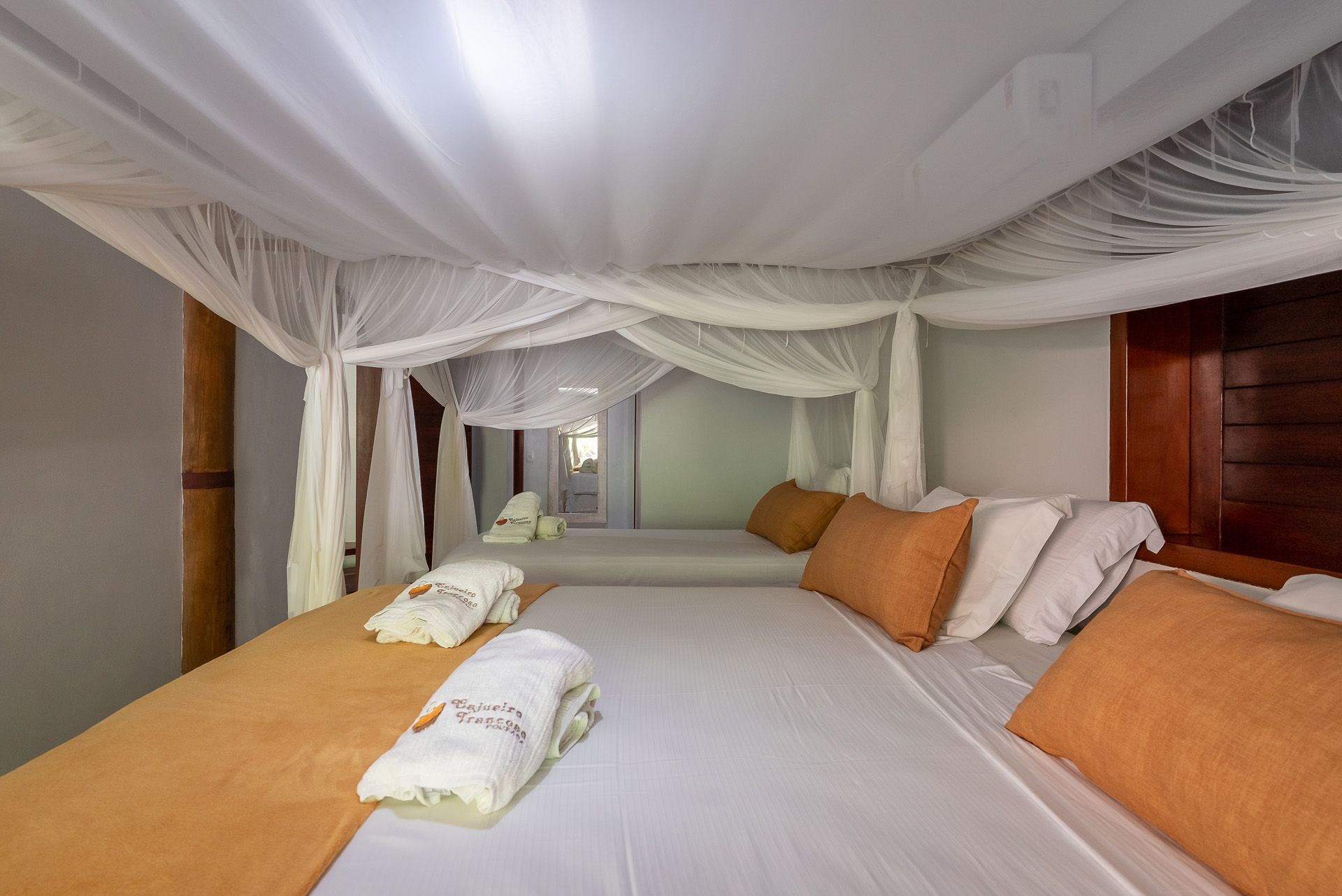 Duas camas com roupa de cama branca, mantas laranja, travesseiros e mosquiteiros em um quarto.