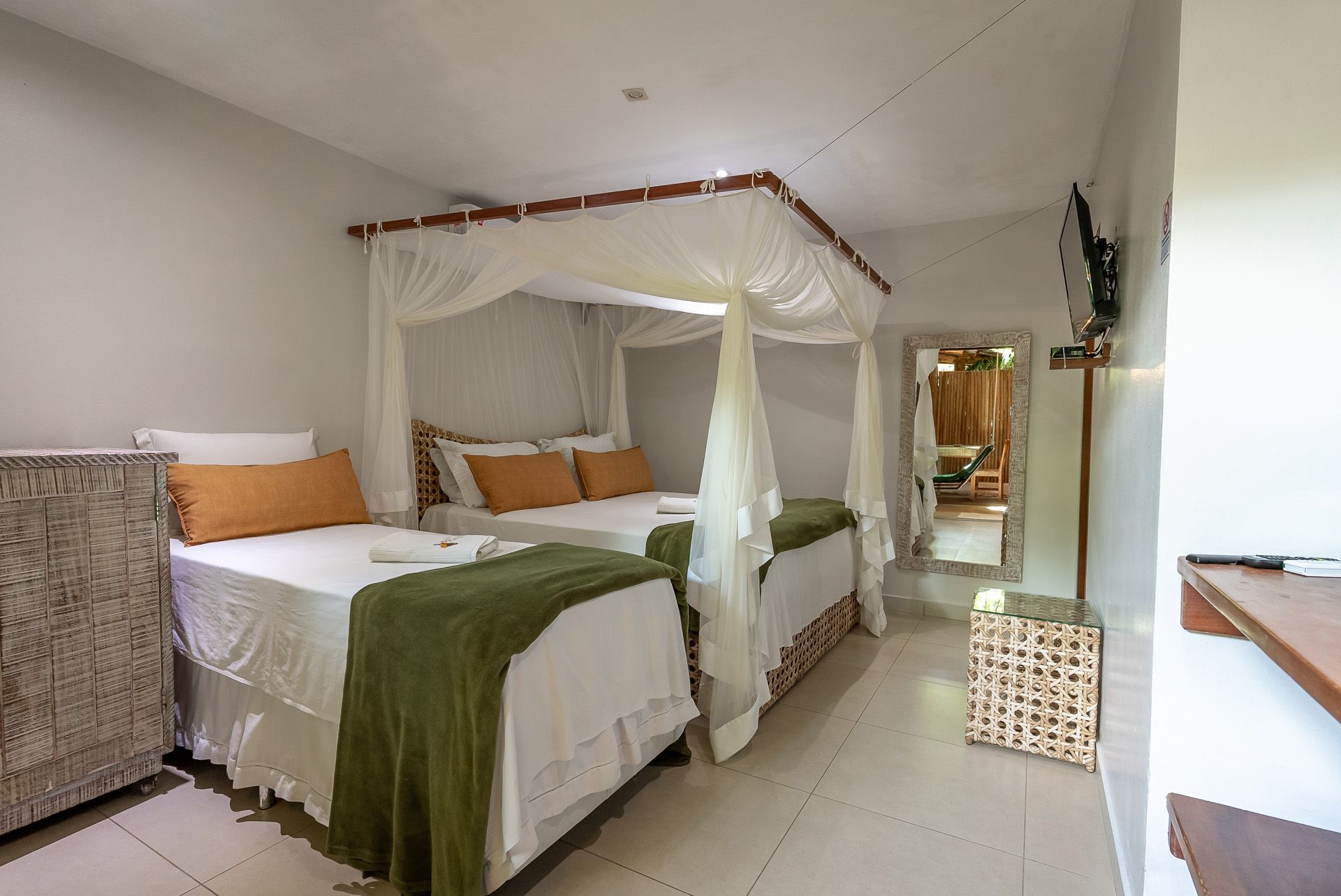 Quarto com duas camas, uma com dossel. Lençóis brancos, almofadas laranja, manta verde, paredes em tons neutros.