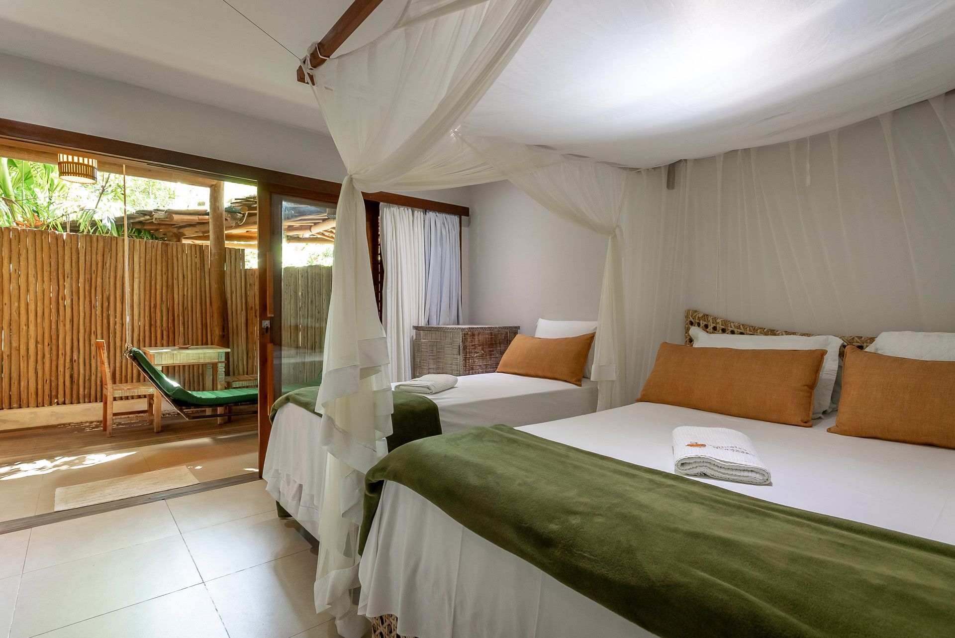 Quarto com duas camas, mosquiteiro, cobertores verdes e acesso a um pequeno pátio.