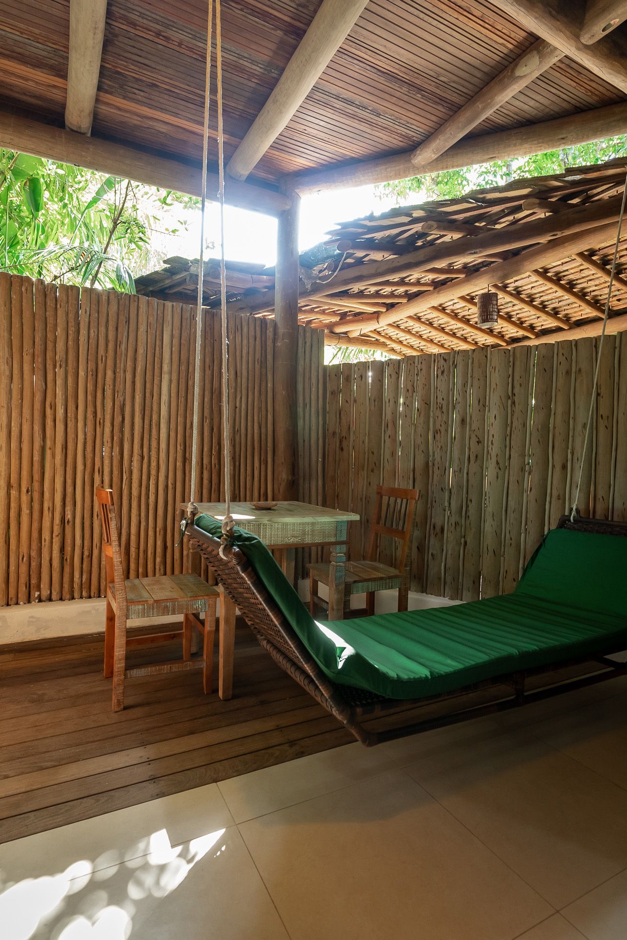 Espaço externo coberto com rede, mesa, cadeiras e paredes de bambu. Colchão verde, teto de madeira.