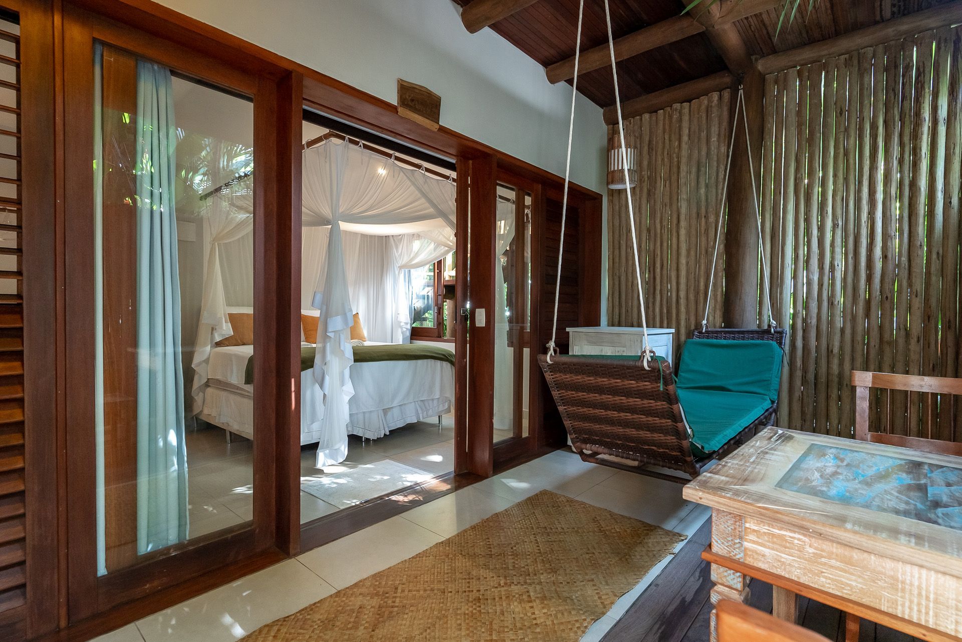 Quarto com cama de dossel visível através de portas de madeira abertas, cadeira de balanço em uma varanda.