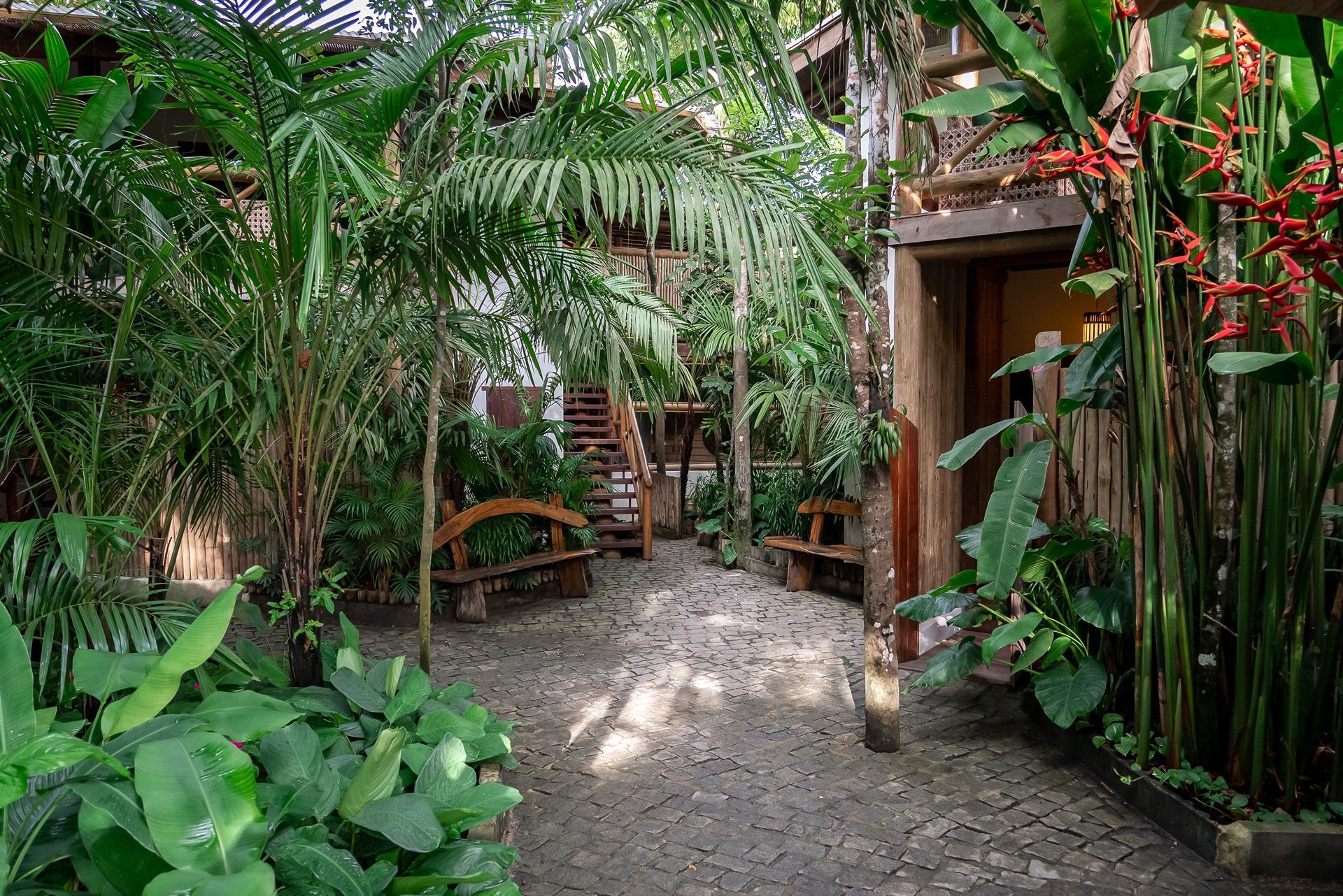Pátio com plantas tropicais exuberantes, caminho de cascalho, bancos de madeira e entradas para o edifício.