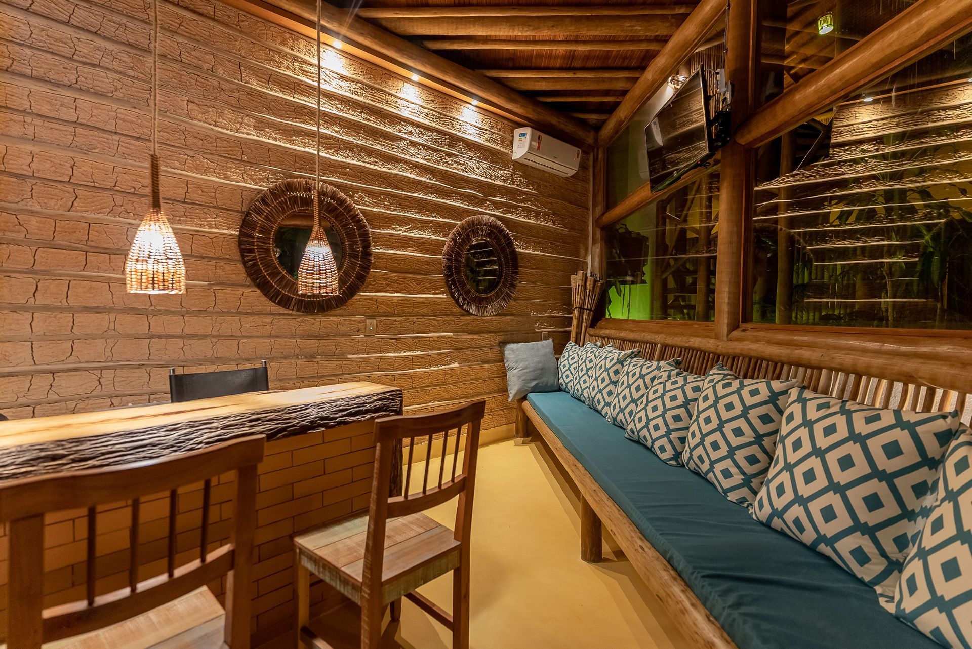 Aconchegante área de estar externa com móveis de madeira, almofadas verde-azuladas, parede de tijolos, espelhos decorativos e luminárias pendentes.