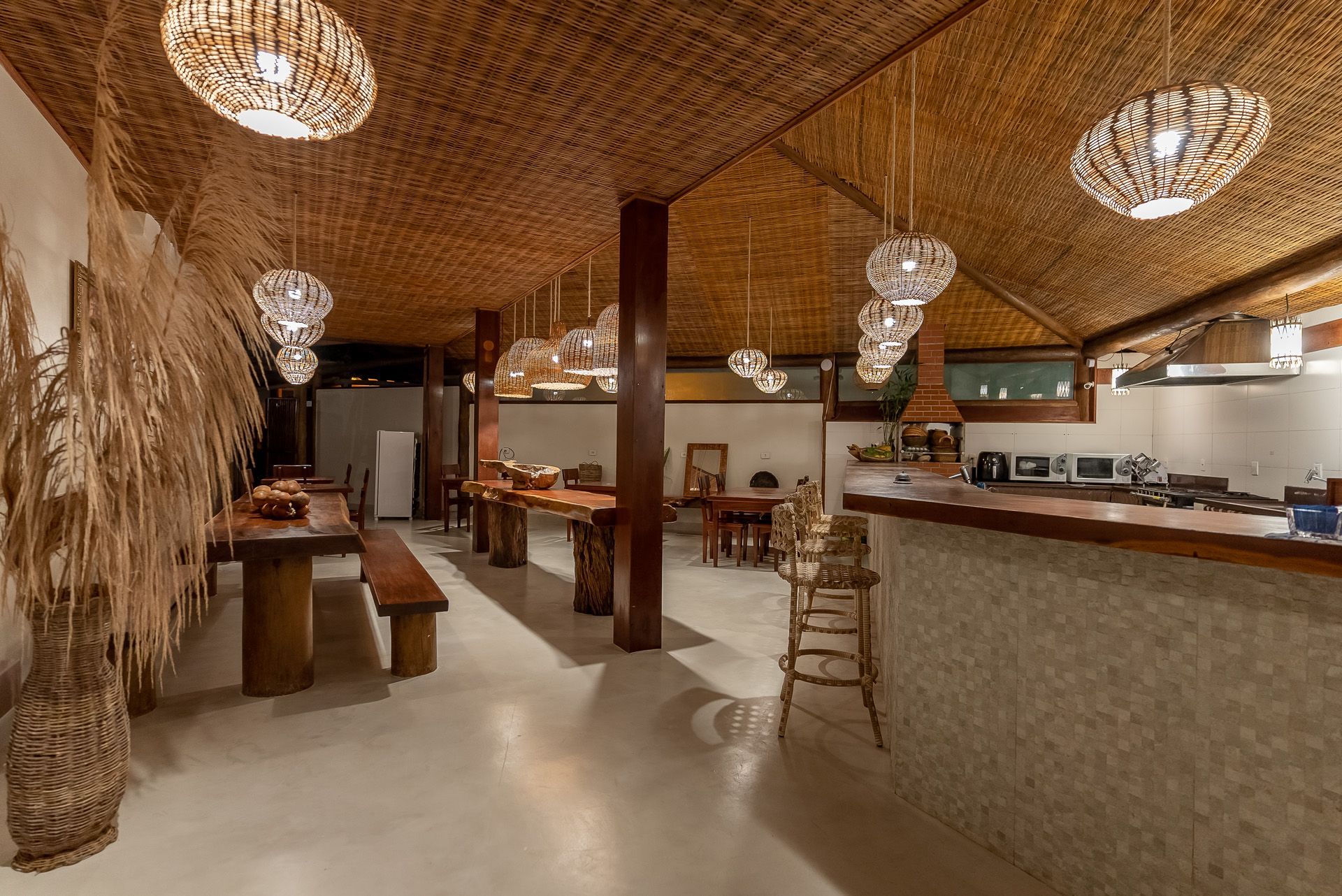 Sala de jantar rústica com mesas e bancos de madeira, teto trançado e luminárias pendentes.