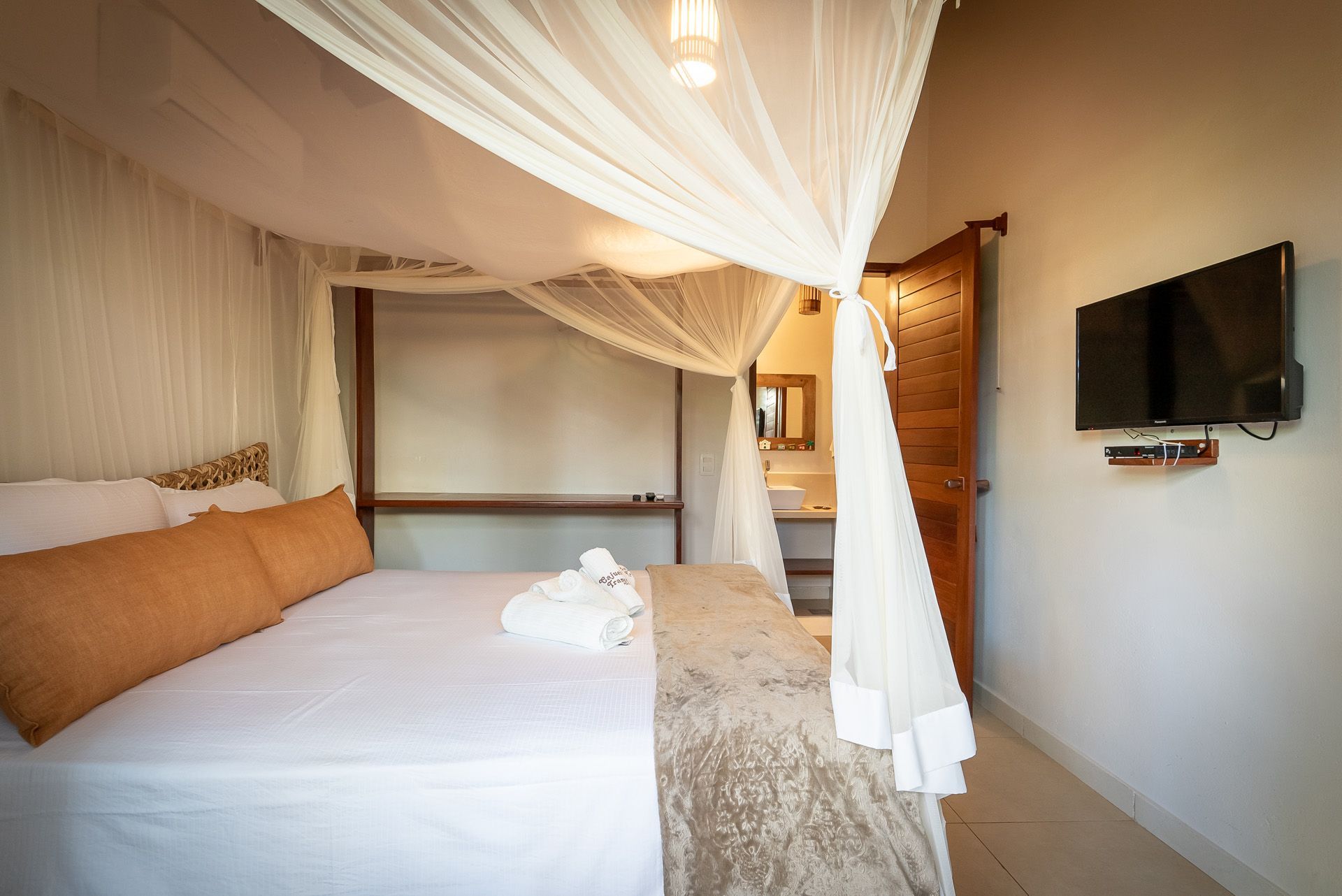 Quarto com cama de dossel, toalhas e TV de parede; porta para um banheiro.