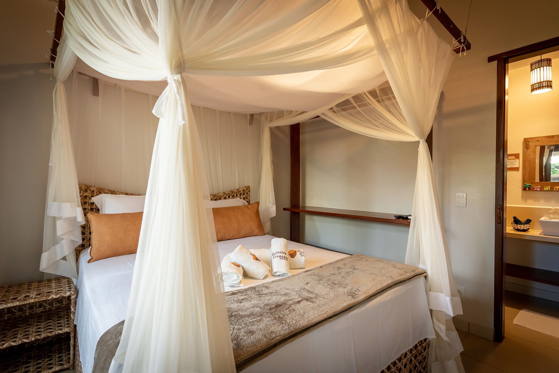 Quarto com cama, dossel, toalhas, almofadas laranja, mesa de cabeceira de vime e banheiro parcialmente visível.