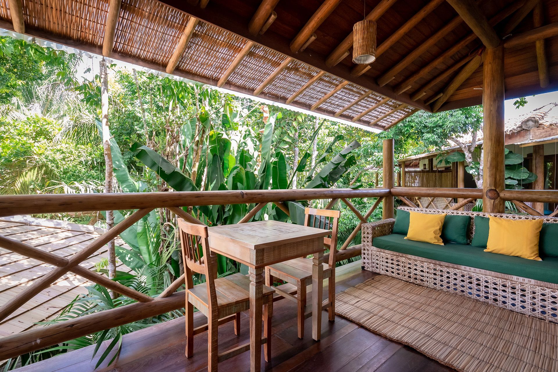 Varanda de madeira com mesa e cadeiras, sofá com almofadas amarelas, com vista para uma folhagem verdejante.