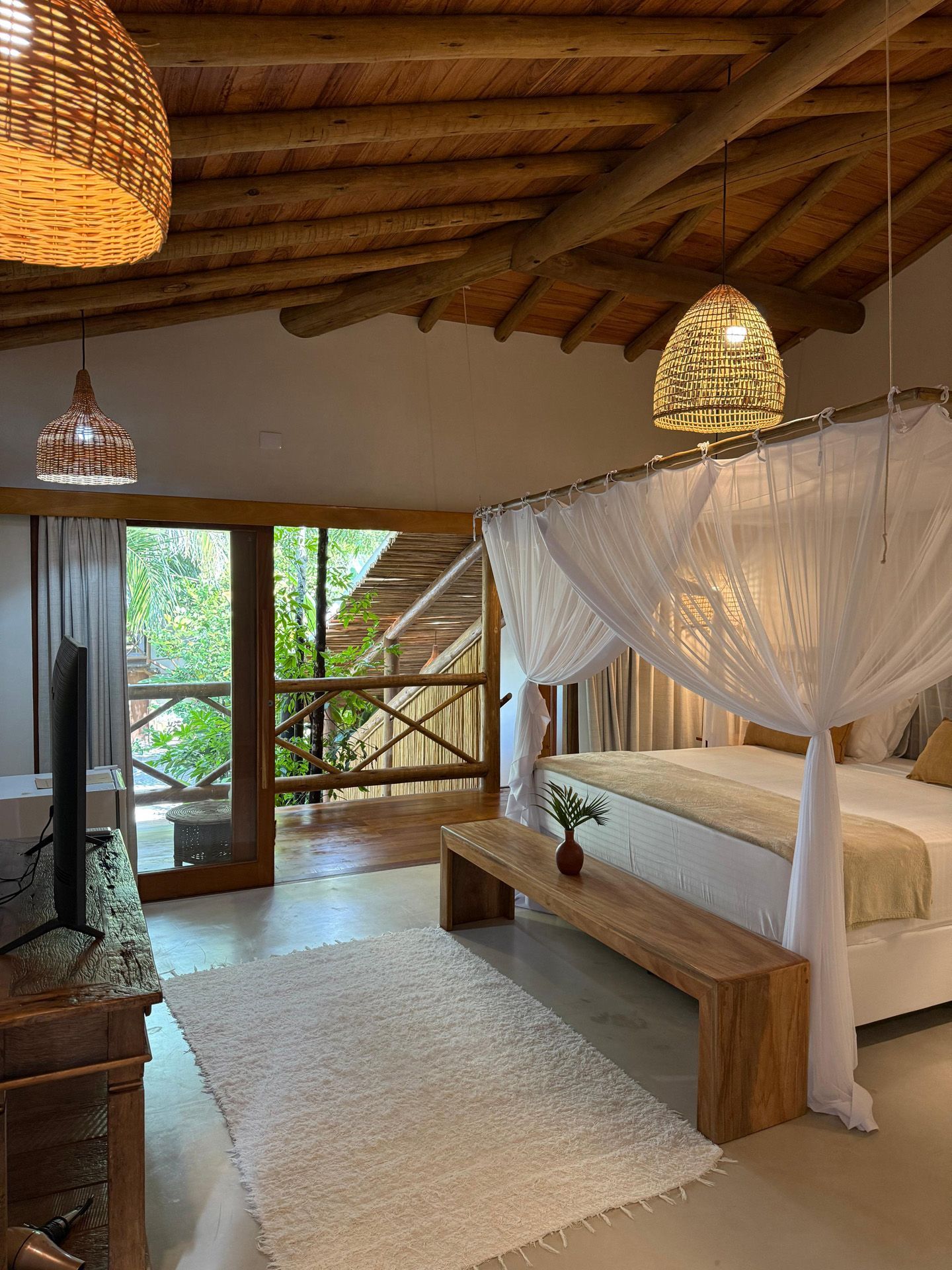 Quarto com cama de dossel, banco de madeira e varanda com vista para uma vegetação exuberante.