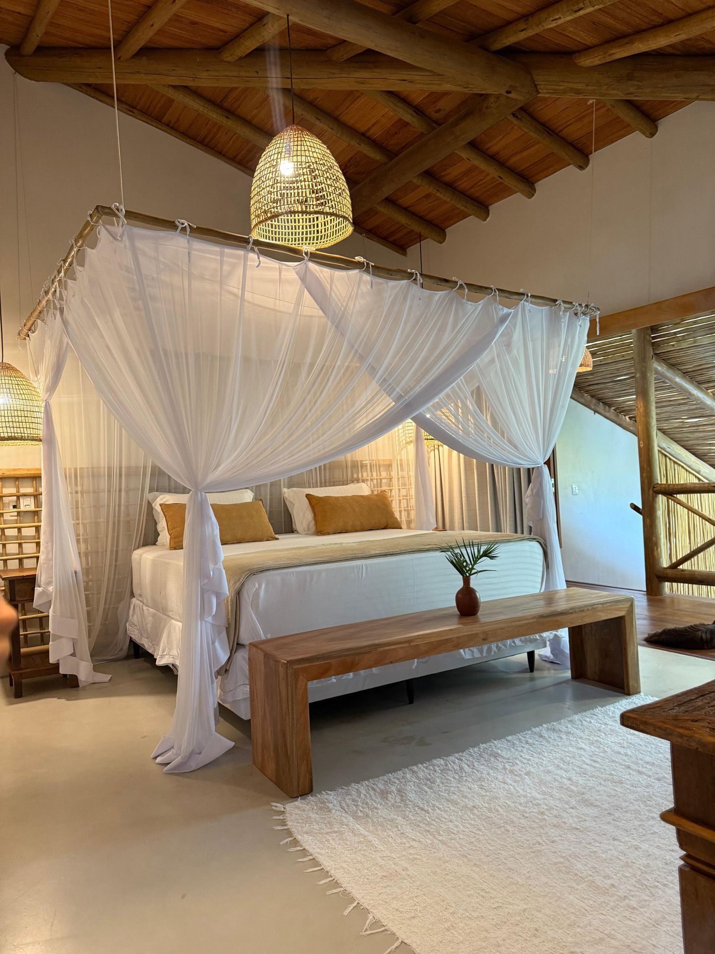 Quarto com cama de dossel, banco, tapete branco e teto de madeira.