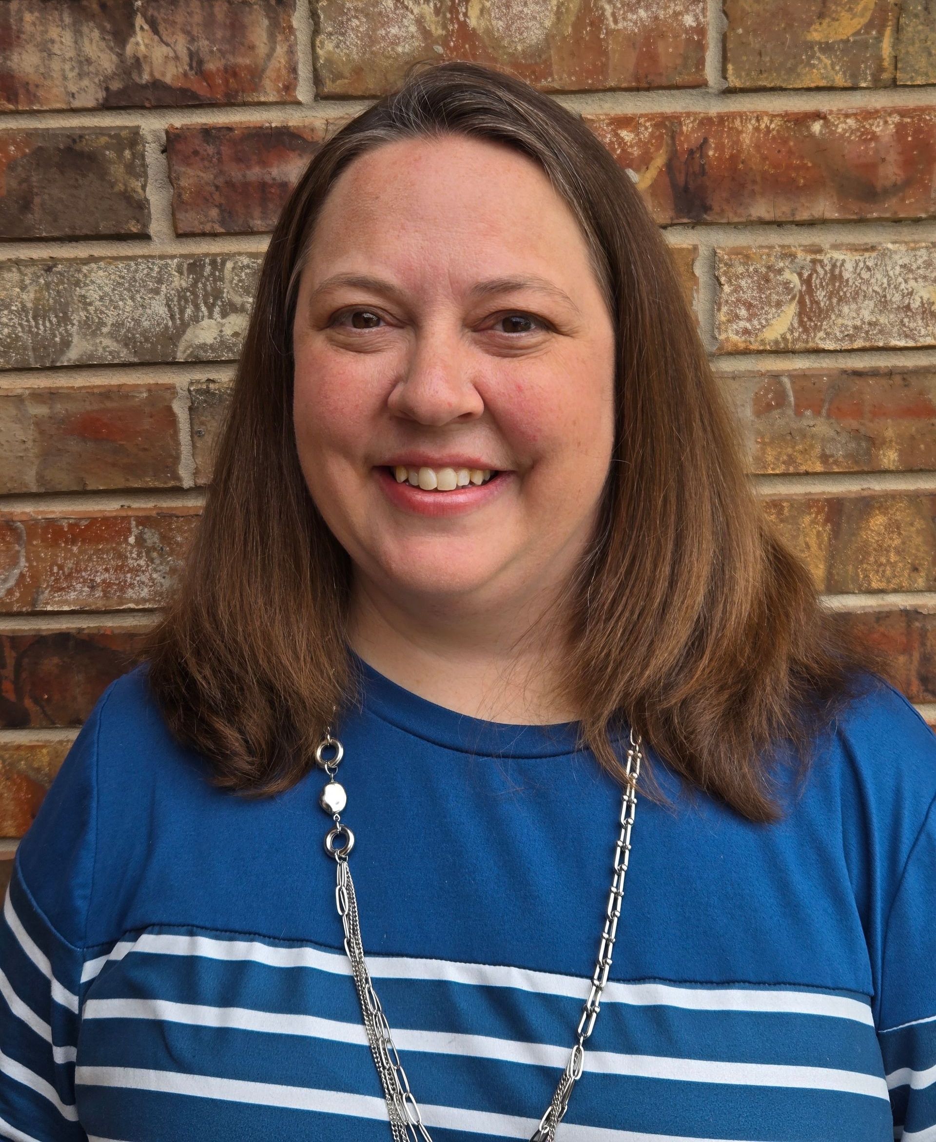 Agent Cara with brown hair smiles, wearing a blue striped shirt and silver necklace, in front of a brick wall.