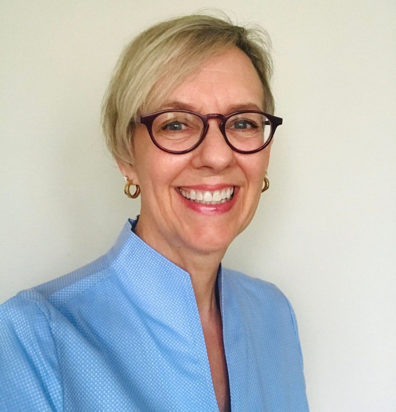 Agent Elly with short blonde hair and glasses smiles at the camera, wearing a blue shirt, indoors.