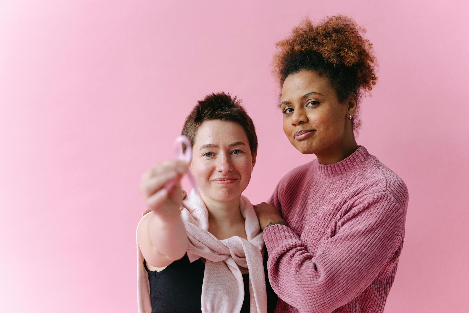 two ladies, a pink backdrop, one holding a pink ribbon, symbolizing breast cancer awareness