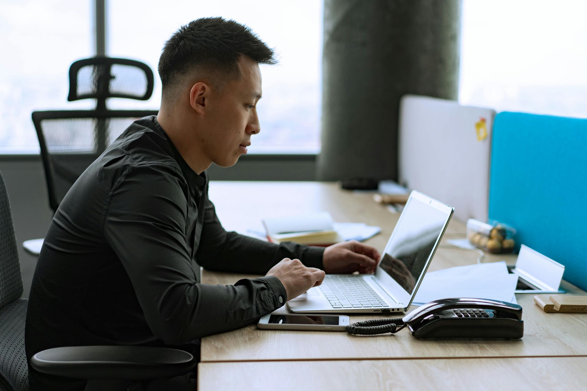 Man in black shirt working on laptop at office desk.