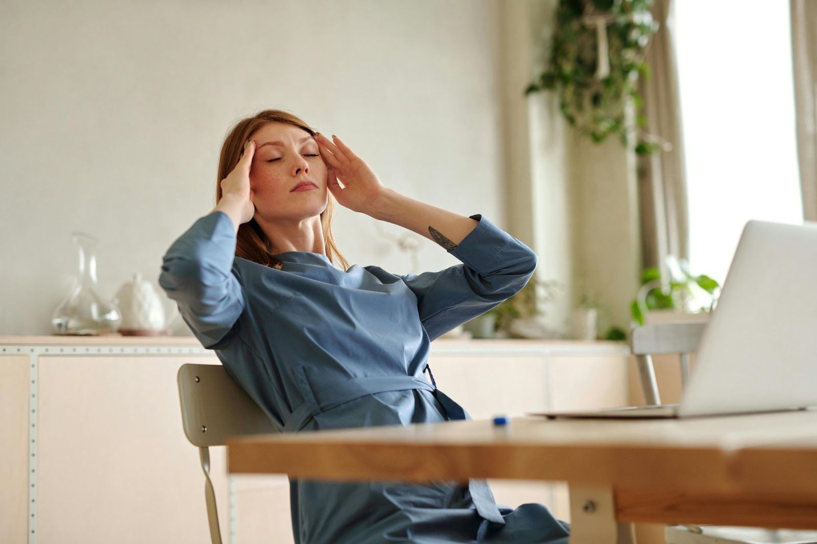 Lady holding her head while closing eyes, lady sitting on the table facing a white laptop