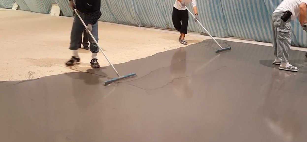 People applying a smooth, gray coating to a floor with long-handled tools in a large indoor space.