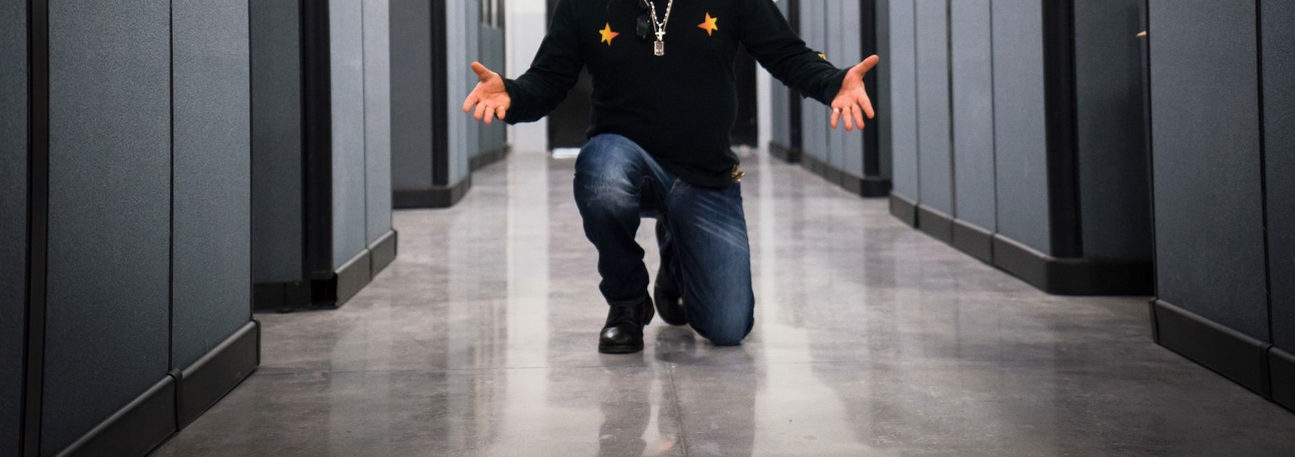 Person kneeling in hallway with hands outstretched. Dark clothes, jeans, reflective floor and walls.