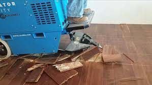 Blue machine removing hardwood floor, pieces of wood scattered on brown floor.
