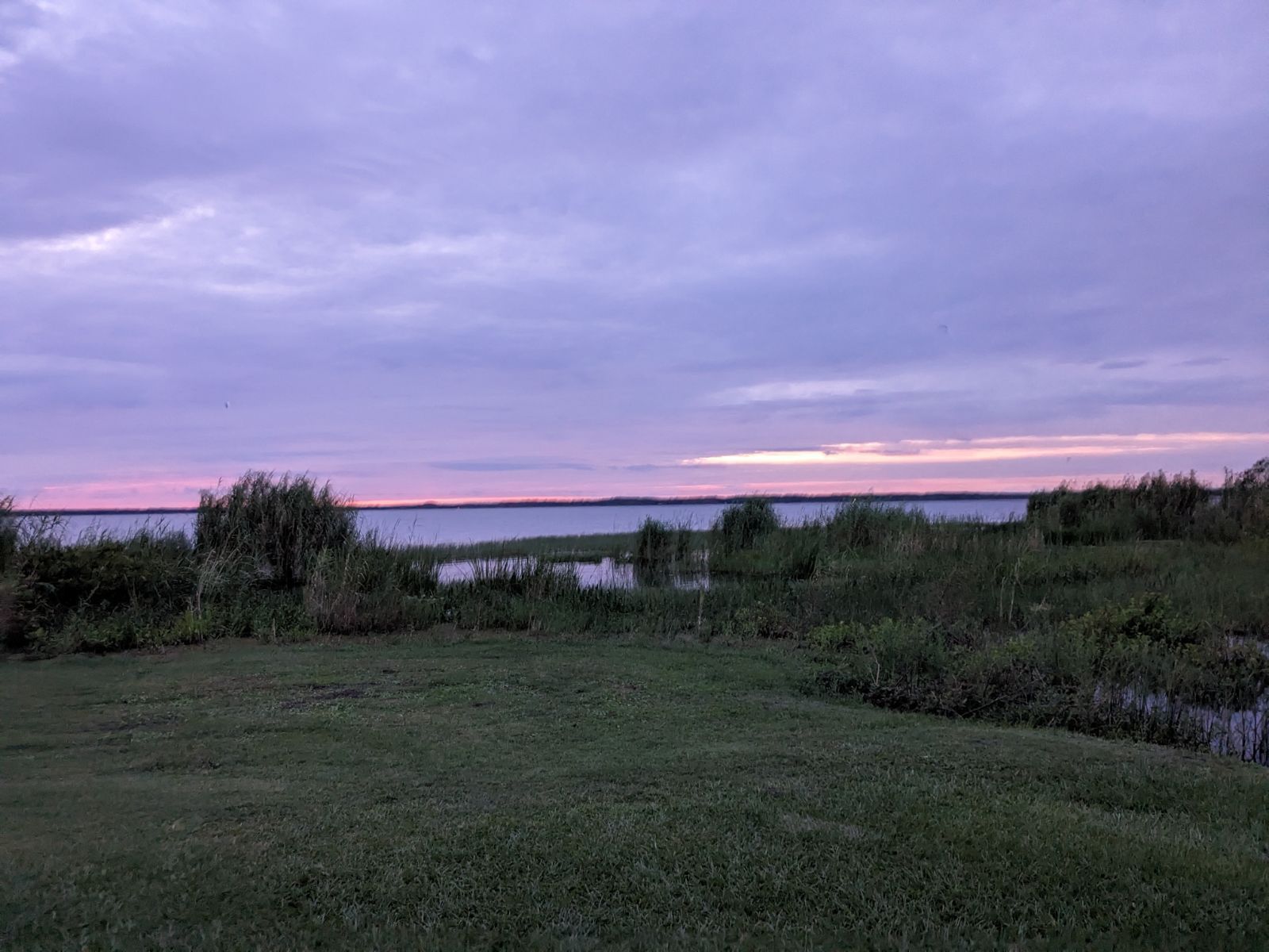 A sunset over a lake with a purple sky and a field in the foreground.