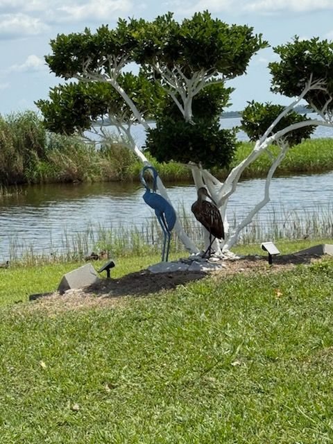 A blue flamingo sits under a tree near a body of water