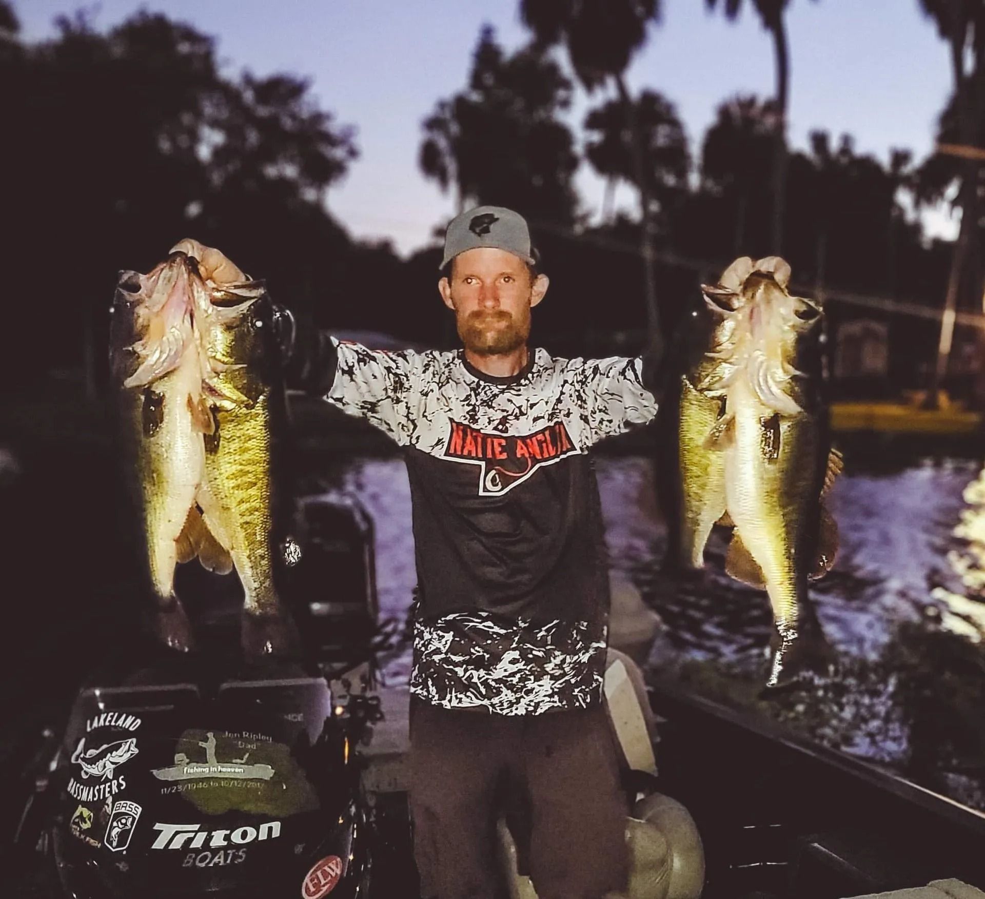 A man holding two fish in front of a boat that says triton