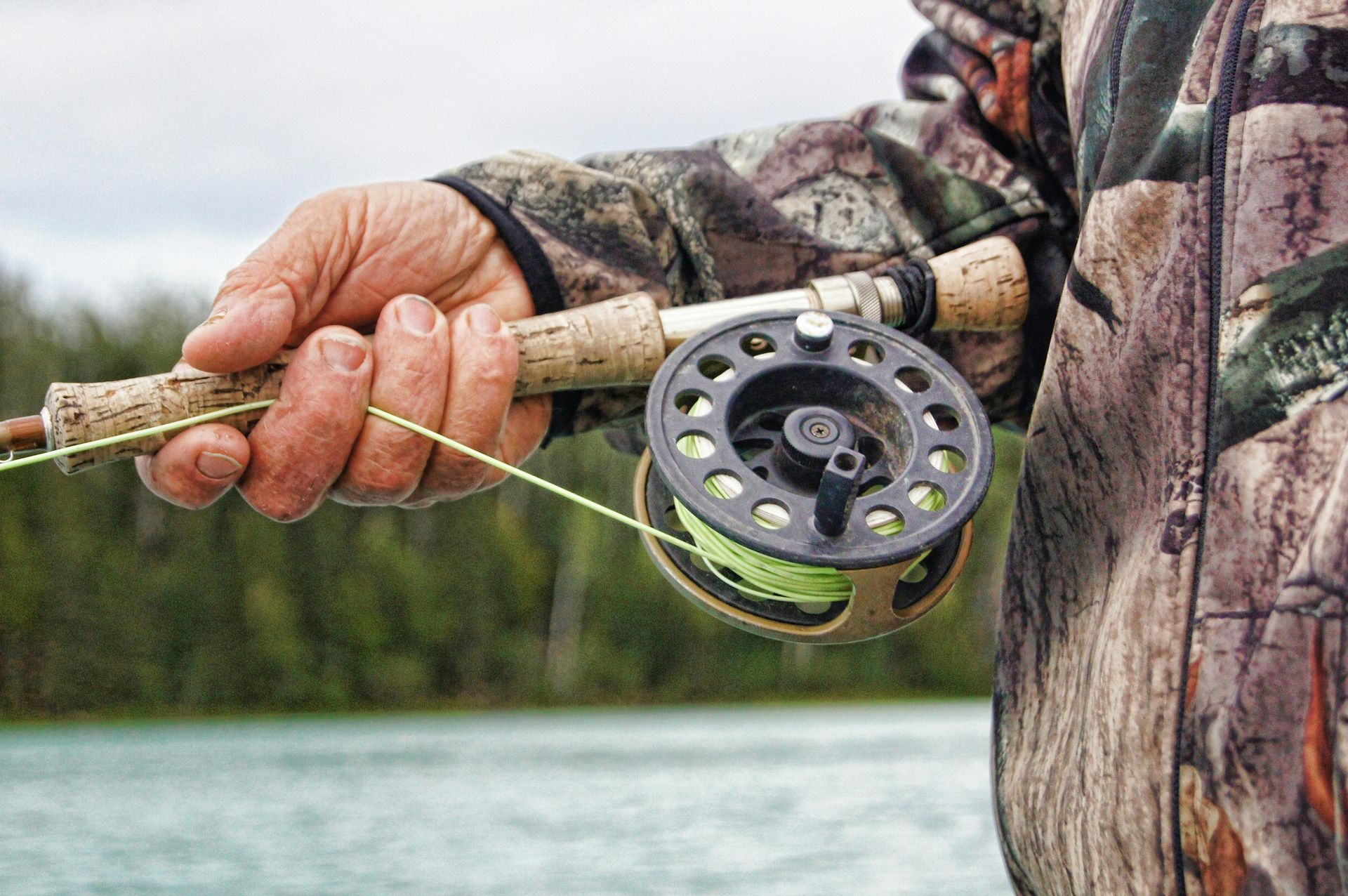 A person's hand holding a fishing rod with a reel and green line, outdoors by water.