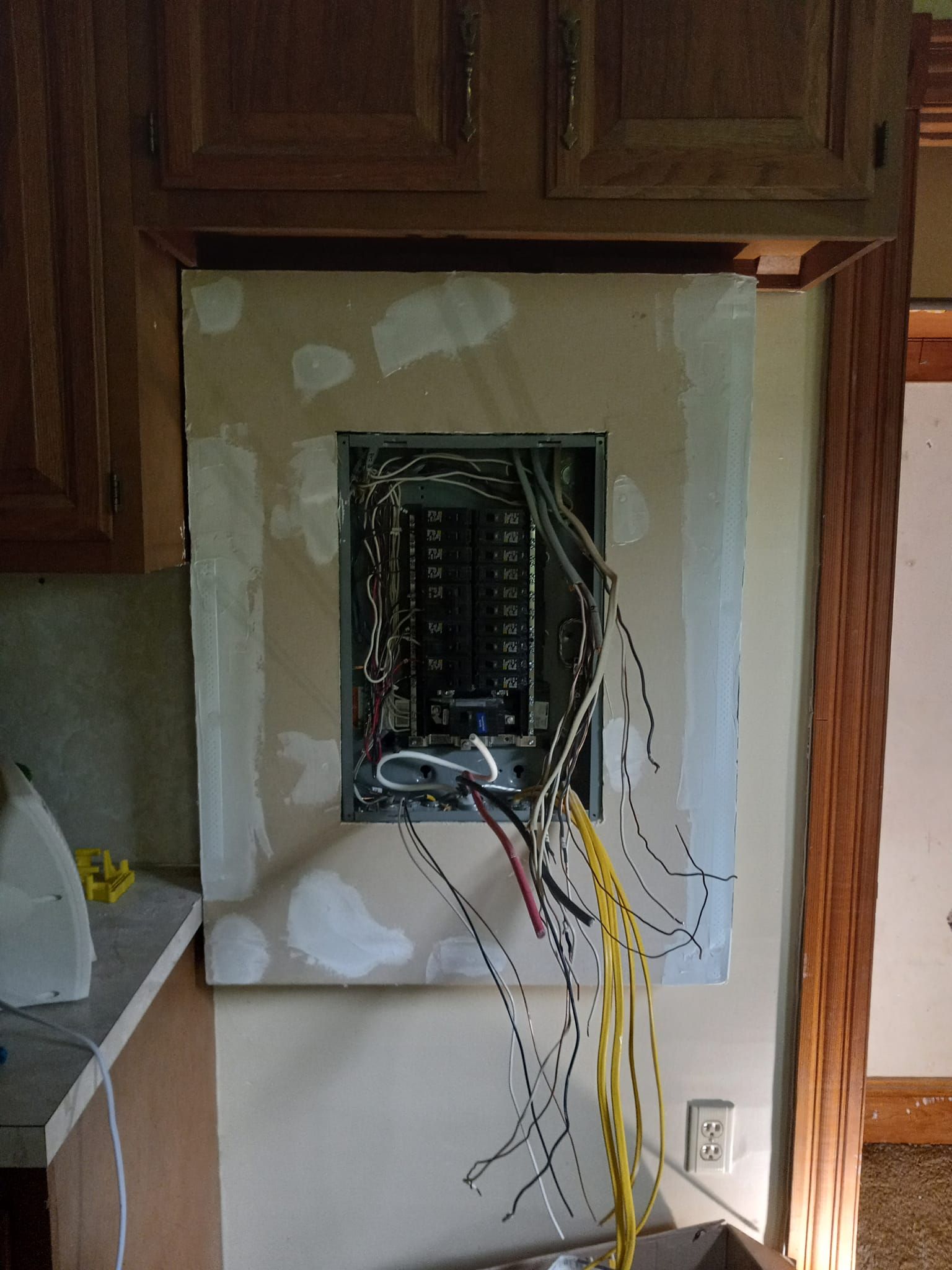 Electrical panel with exposed wires, drywall surround, installed in a kitchen cabinet.