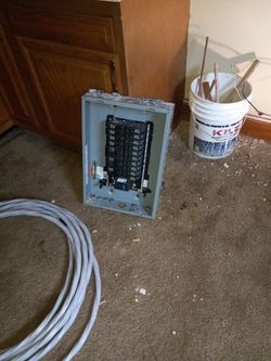 Electrical panel on carpet with wire coil, paint bucket, and wood scraps.