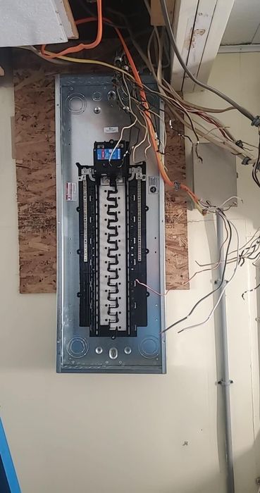 Electrical panel with numerous wires protruding from the top, mounted on a wooden board against a wall.