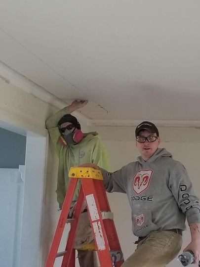 Two people on a ladder working on ceiling trim. One wears a respirator, the other safety glasses.
