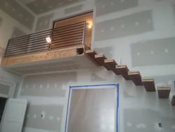 Floating staircase leading to a loft with horizontal metal railing. Interior construction setting, unfinished drywall.