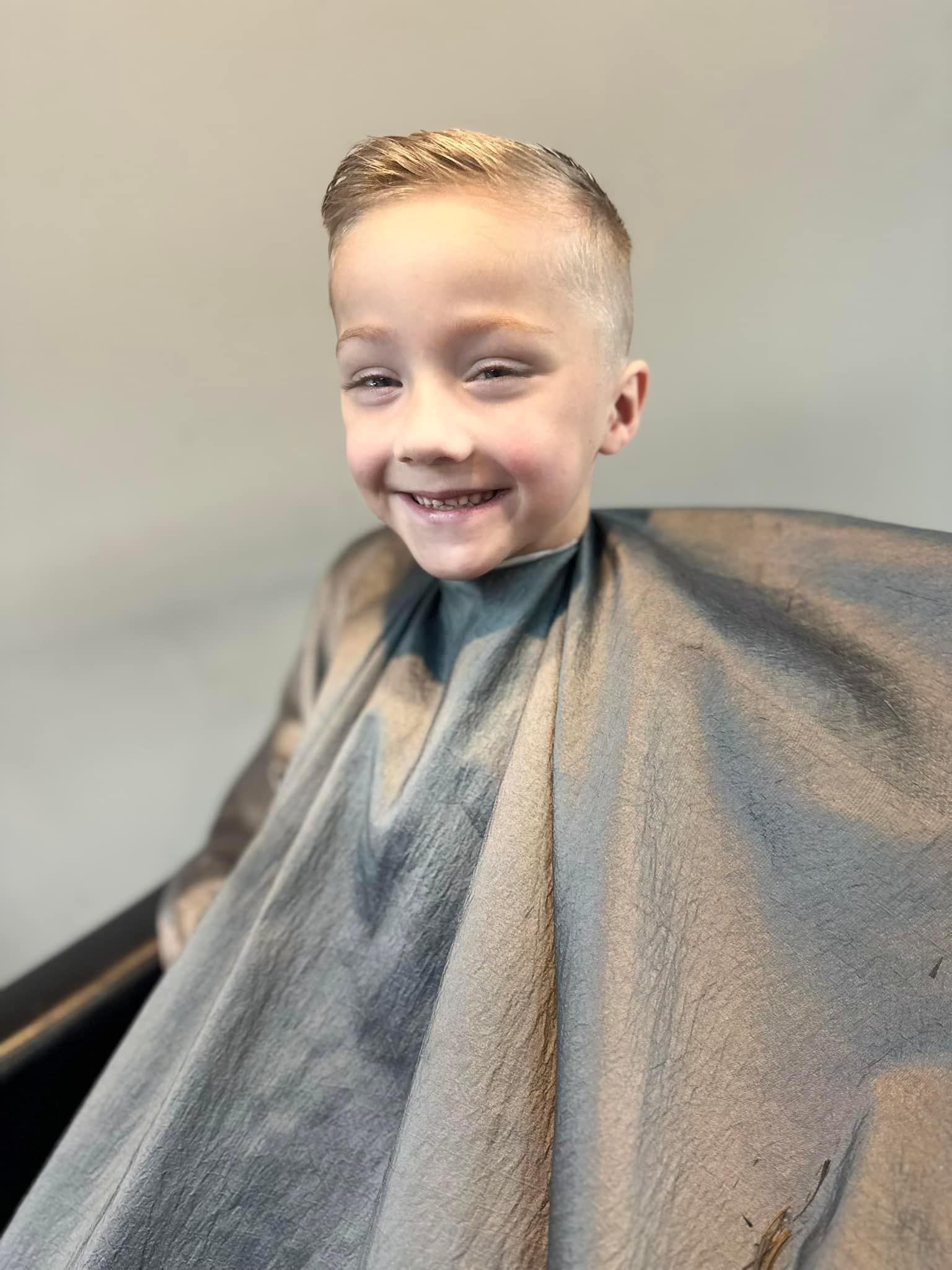 Smiling child with a fresh haircut, wearing a gray barber cape in a salon chair.