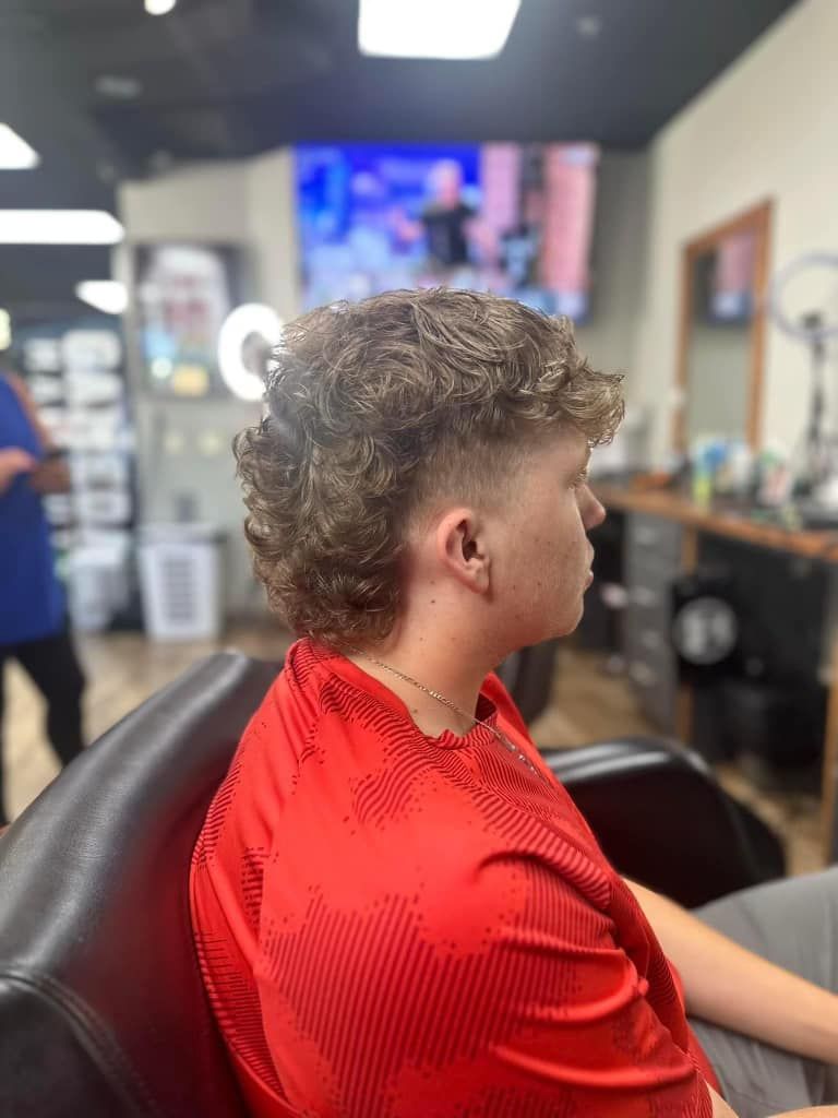Person in a red cape sits in a barber chair, side profile, inside a barbershop.