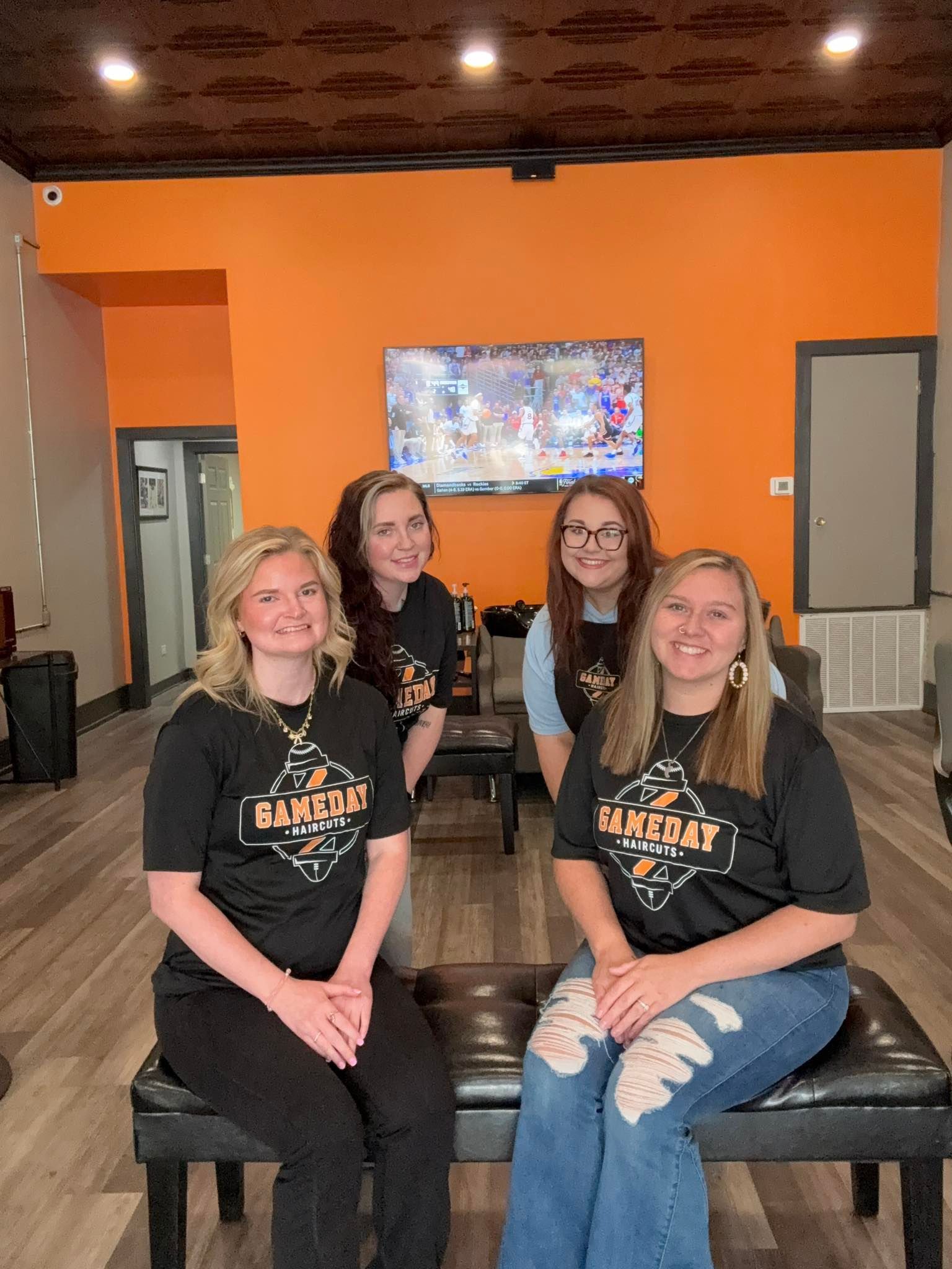 Four people in black shirts pose in an orange-walled room with a TV. Two sit on a bench, two stand.