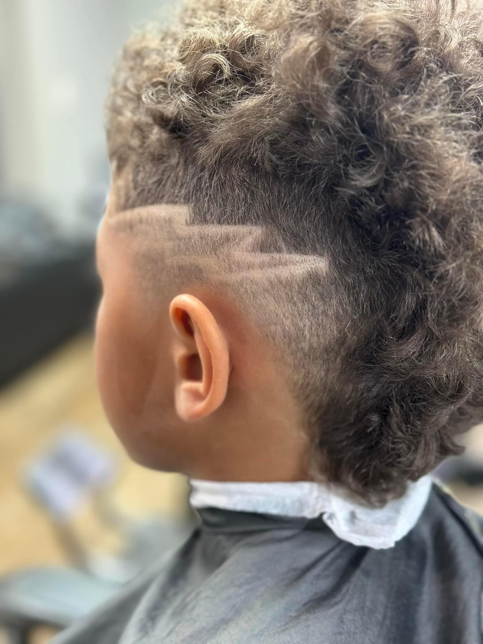 Side view of a child with curly hair and a shaved design haircut, seated in a barber cape.