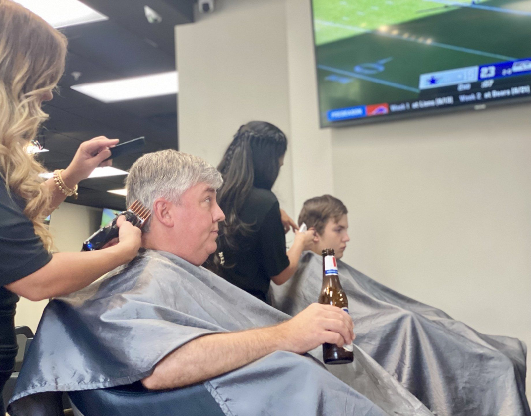 Man getting a haircut while holding a beer in a salon; another person getting their hair cut; TV in background.