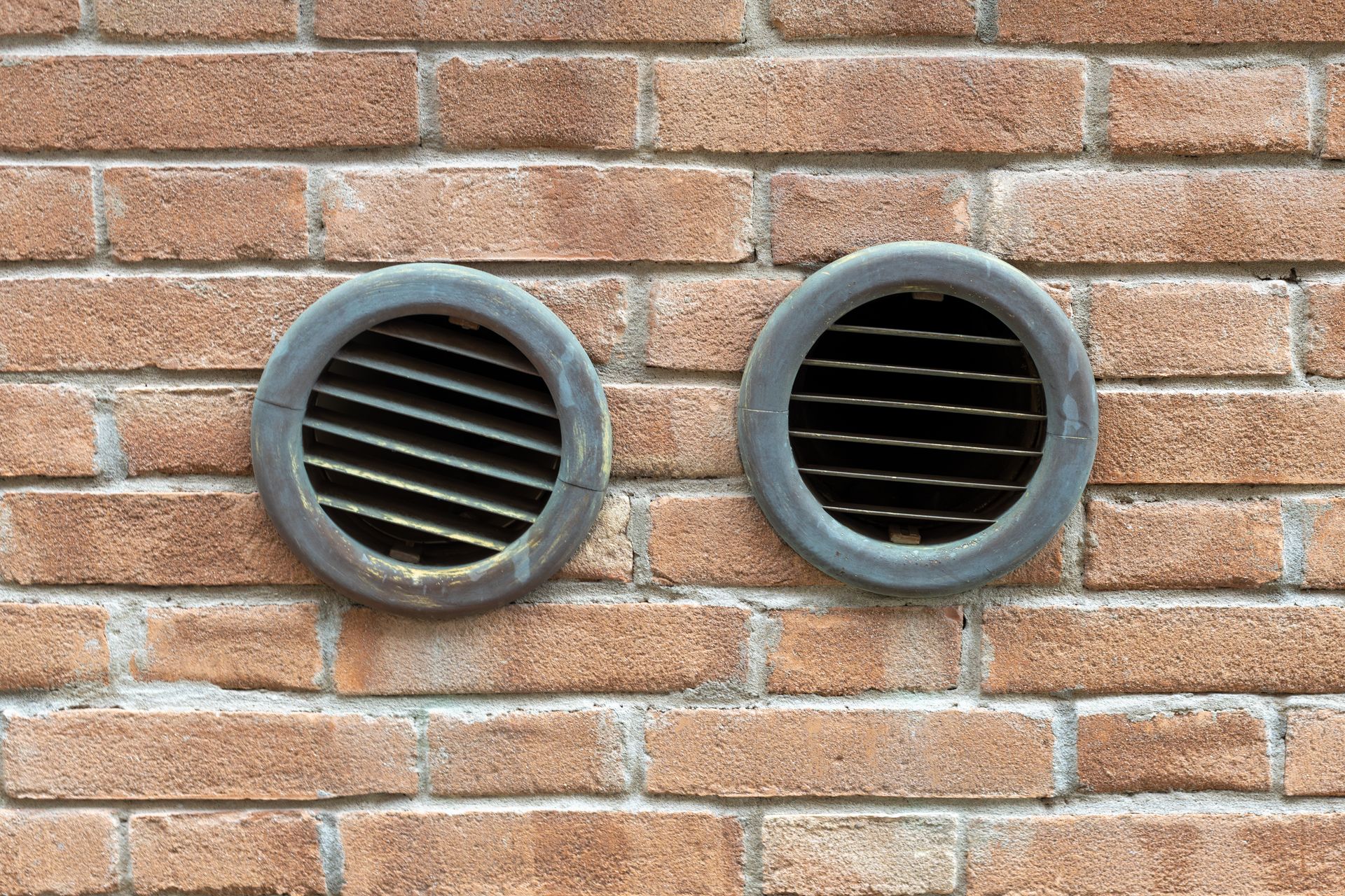 Two circular, grey metal ventilation grilles mounted side-by-side on a textured brown brick wall.