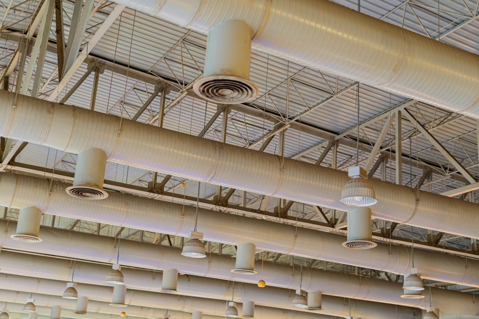 Industrial ceiling featuring white ductwork, circular air vents, and suspended lights against a metal truss structure.